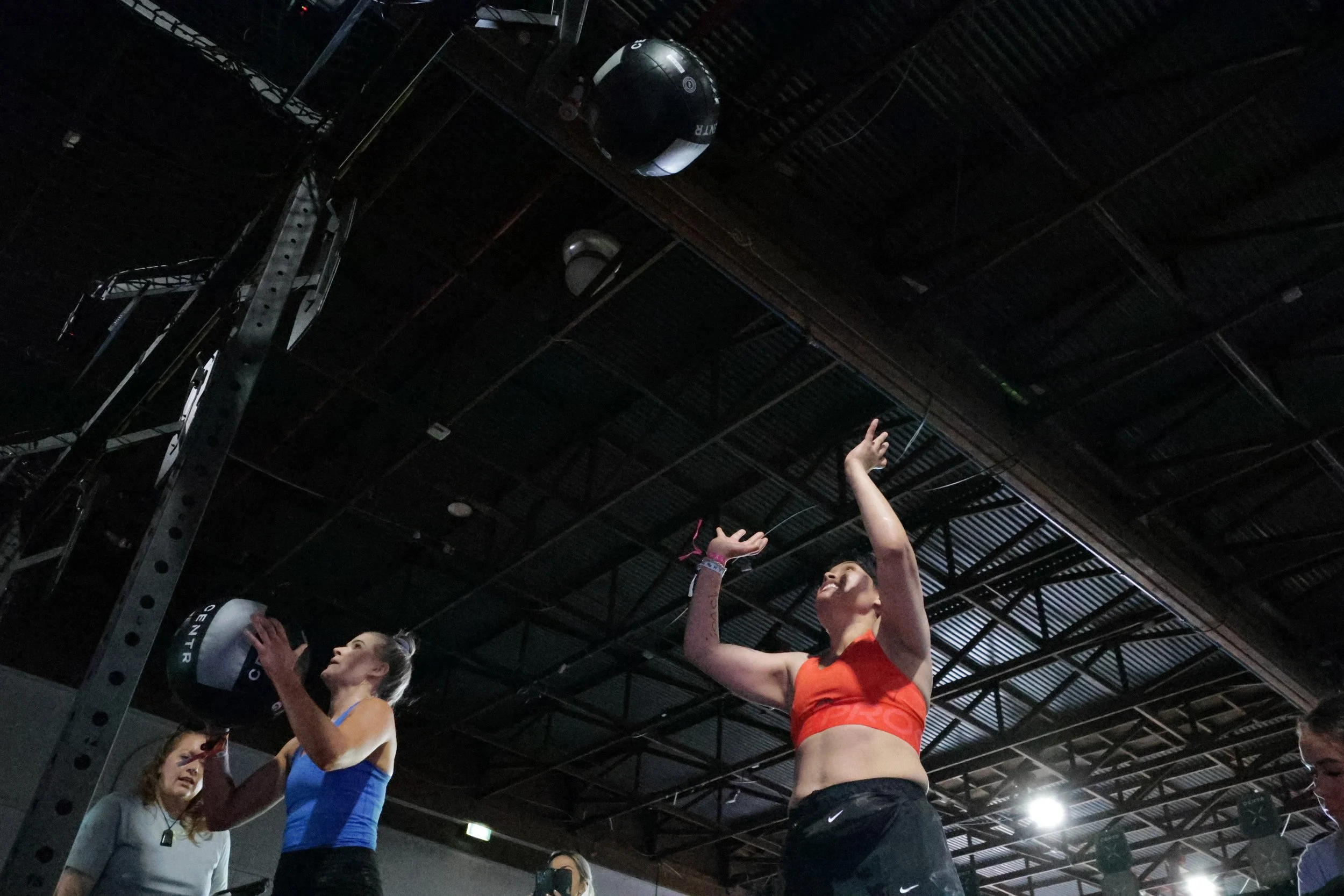 Group of female fitness instructors or athletes in workout clothing, standing in a gym or fitness studio with a high ceiling and exposed beams, some raising their arms and engaging with each other.