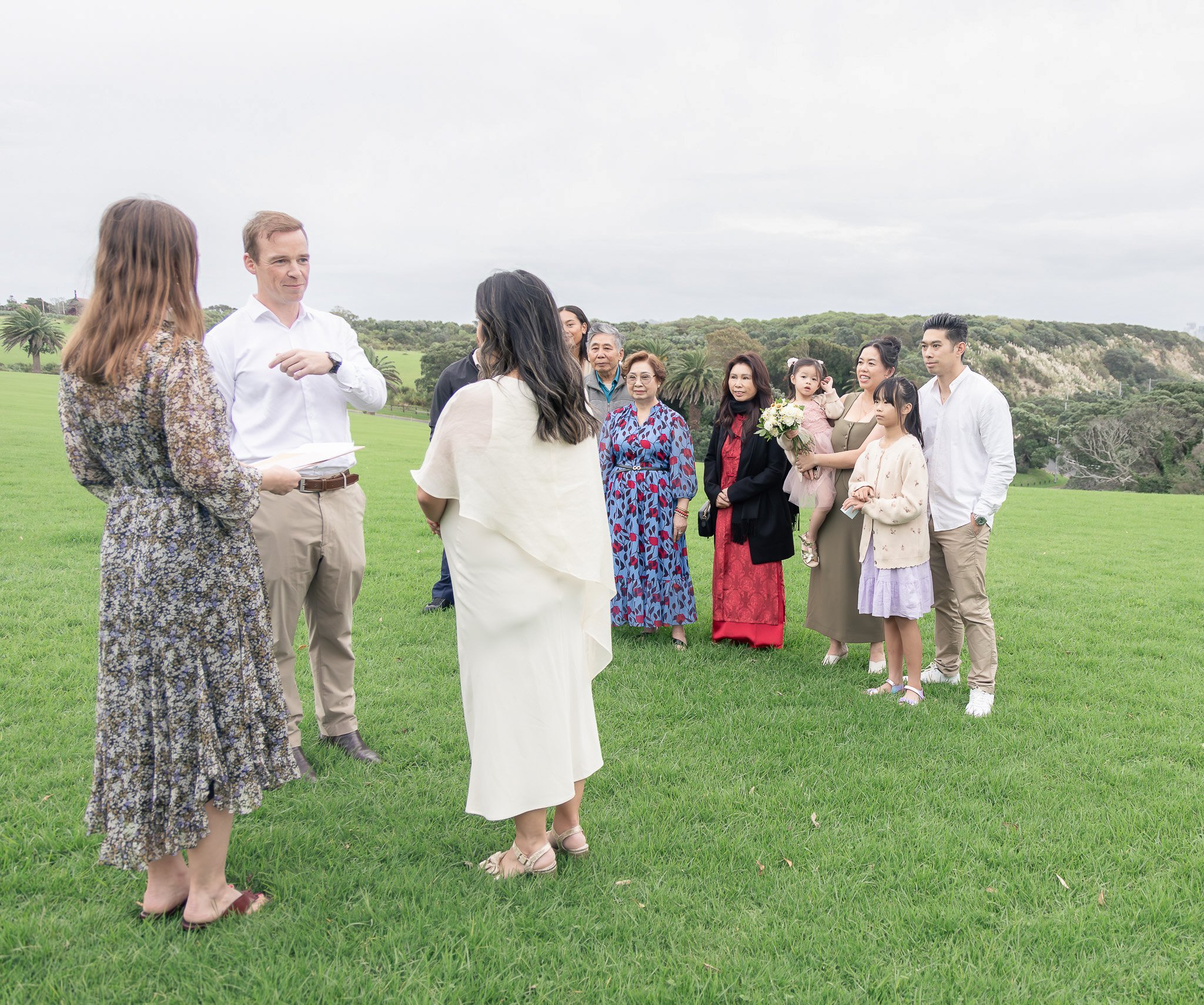 A group of people gathered outdoors on a grassy area, with a cloudy sky and trees in the background. Some individuals are talking, while a woman holds a bouquet of flowers.