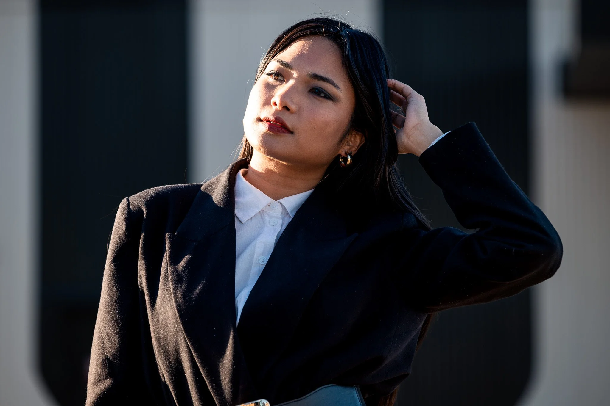A young woman with dark hair and hoop earrings, wearing a white shirt and black blazer, posing outdoors with one hand in her hair and sunlight on her face.