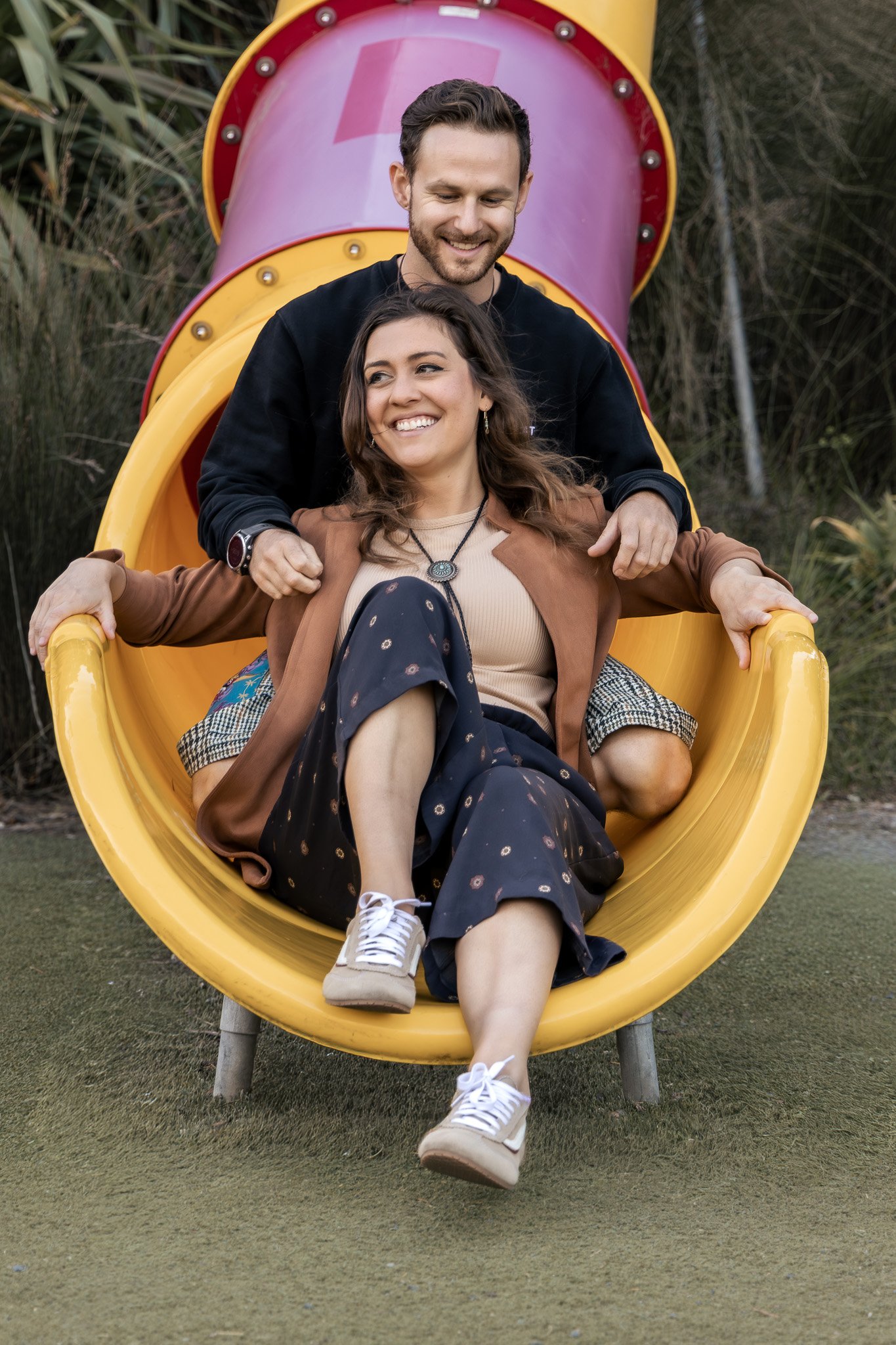 A smiling man and woman on a playground slide, with the man standing behind the woman holding her shoulders, and the woman sitting at the bottom of the slide smiling with crossed legs, surrounded by trees and grass.