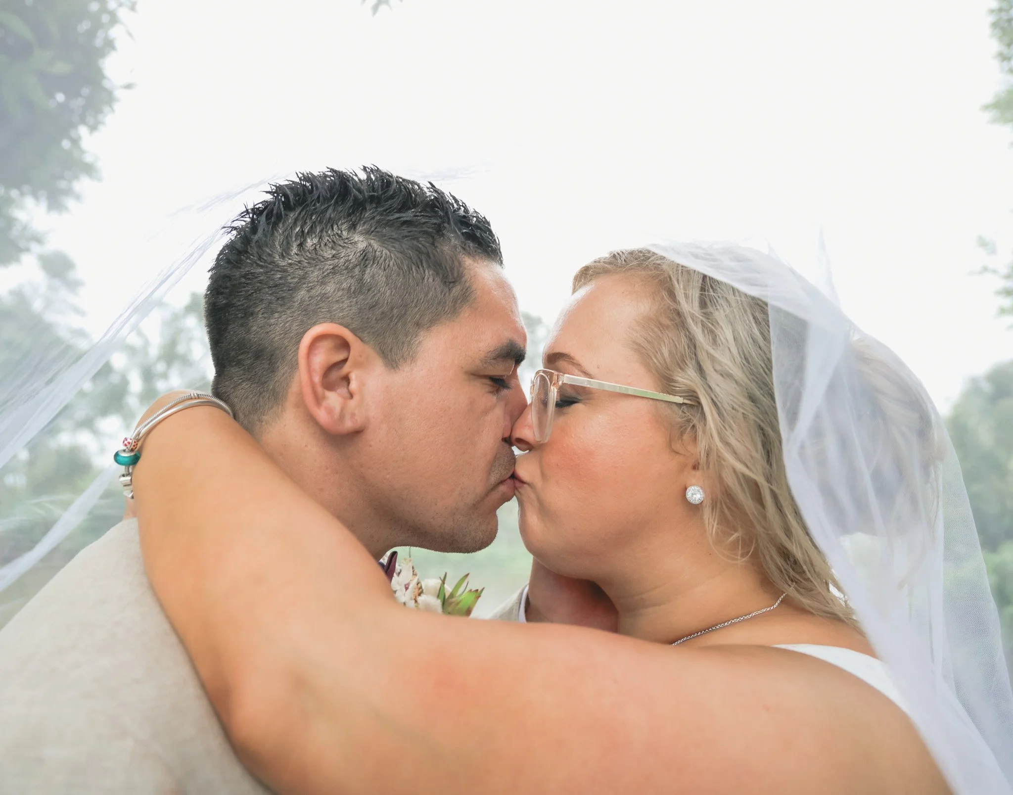 A couple is kissing outdoors under a veil, with trees in the background.