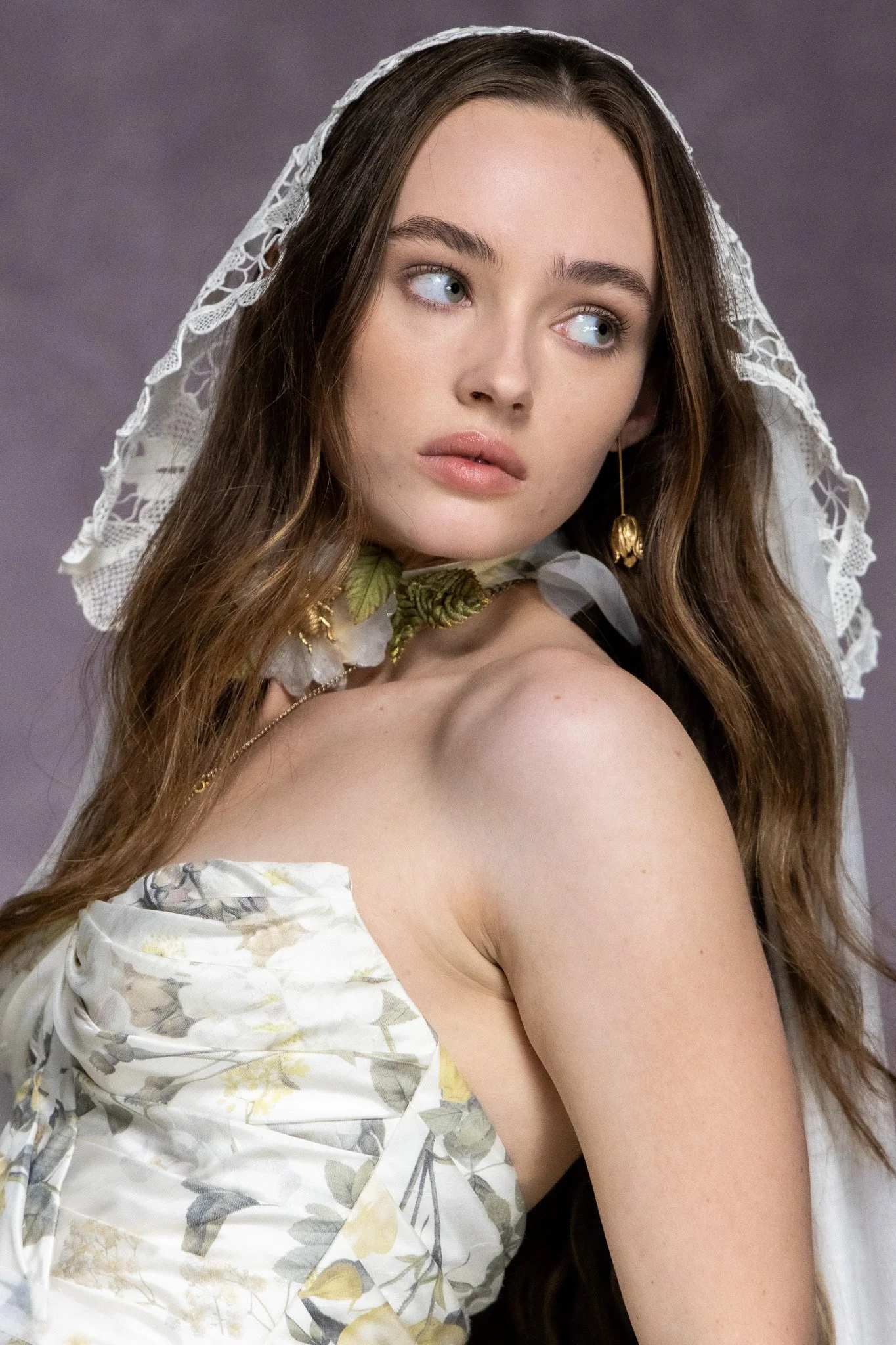 A young woman with long brown hair, blue eyes, and fair skin wearing a floral dress with a strap that exposes her shoulder. She has a lace veil draped over her head and a floral necklace, with a neutral facial expression, looking slightly to her left