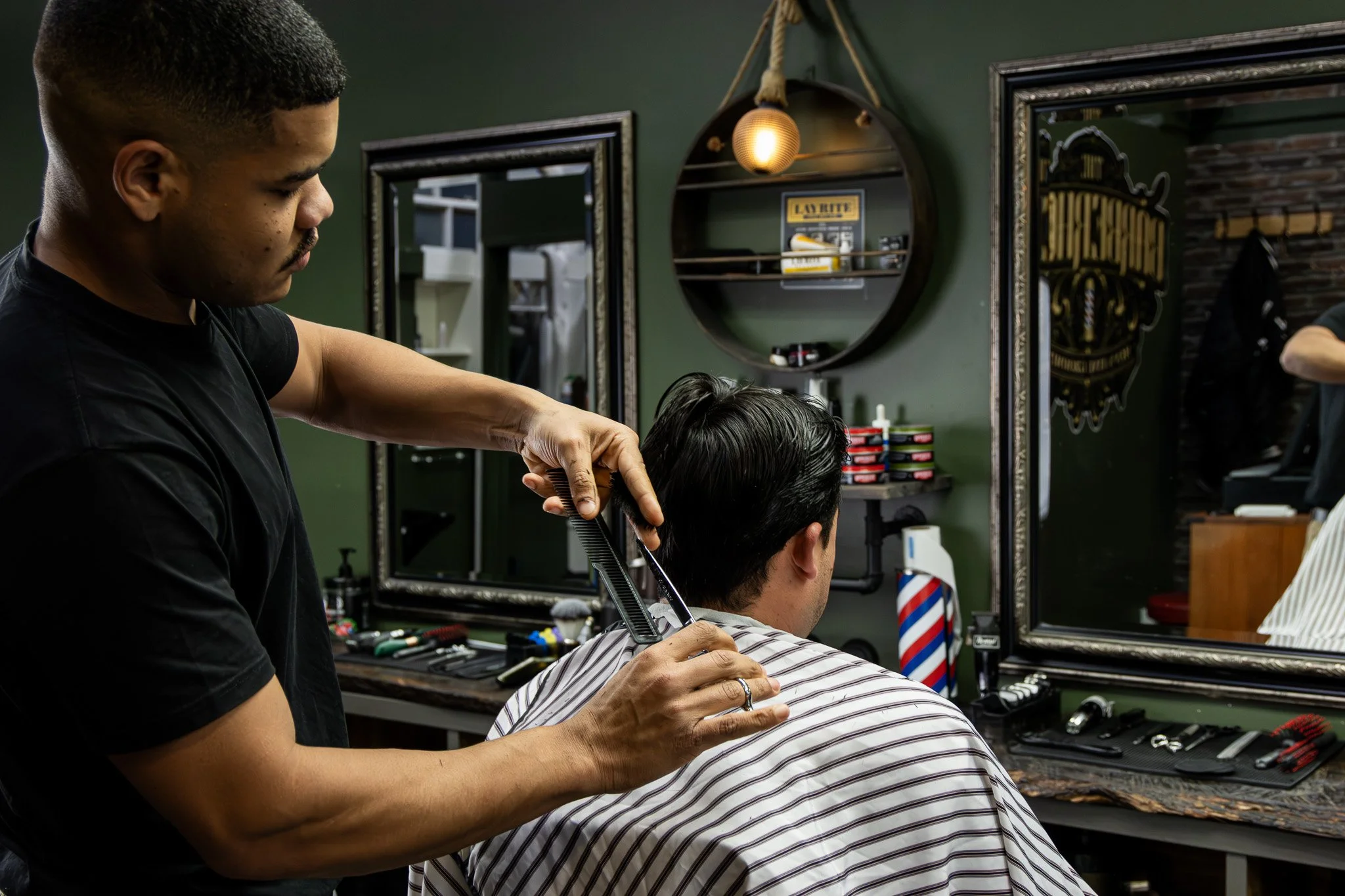 Barber trimming a man's hair in a barbershop with green walls and mirrors.
