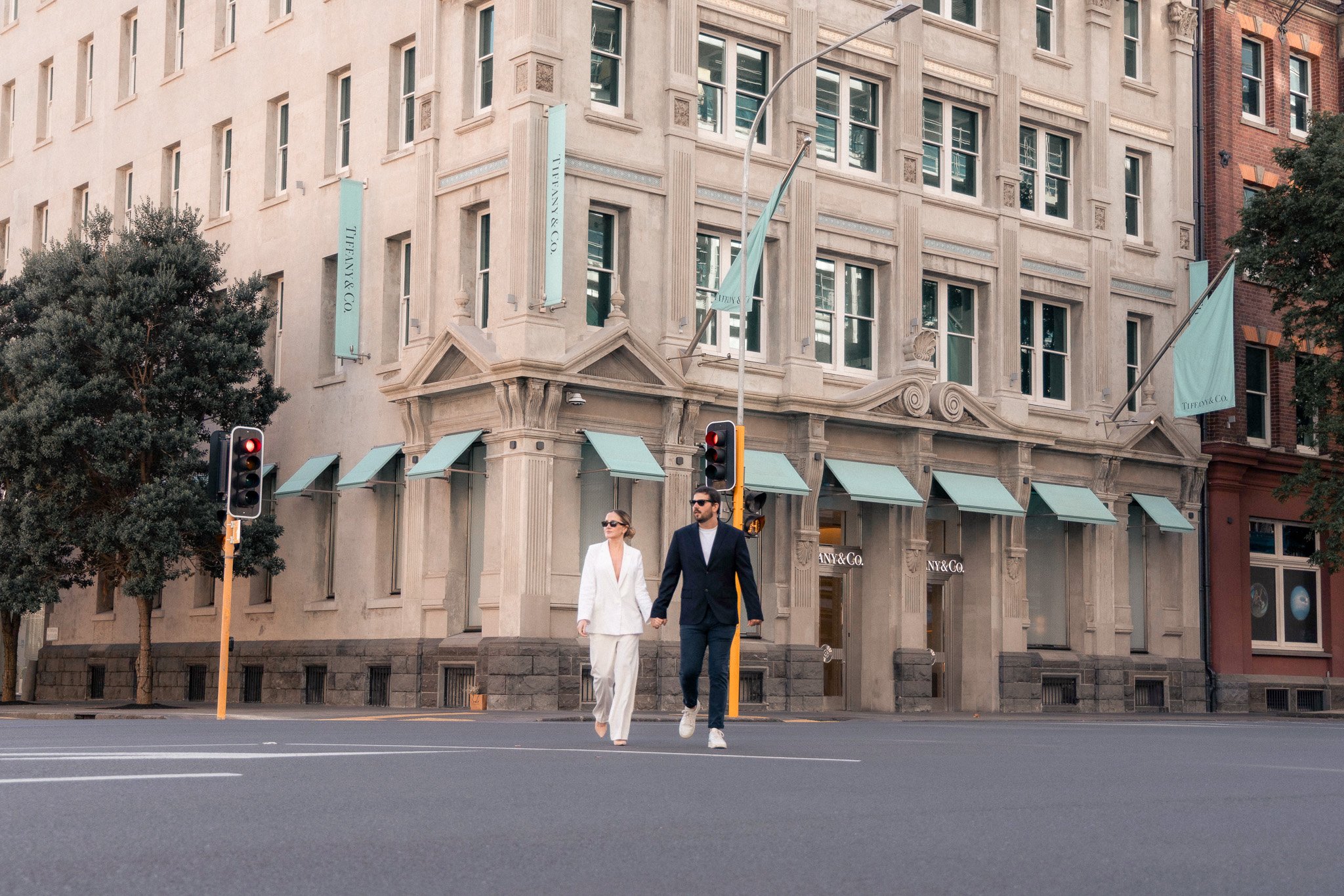 A man and woman holding hands crossing the street in front of a Tiffany & Co. building with beige architecture and teal banners, traffic lights, and trees lining the sidewalk.