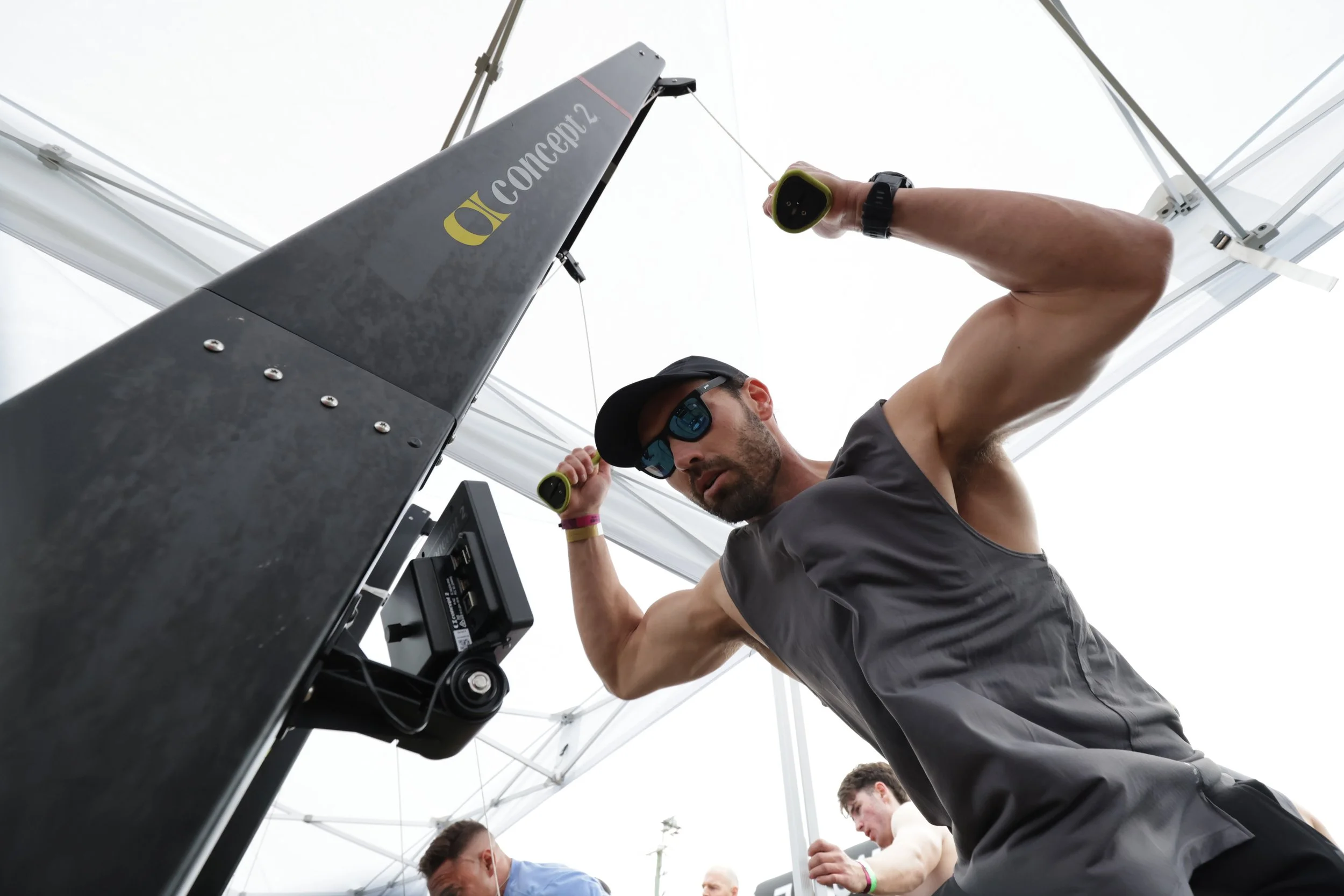A man with sunglasses and a black watch is adjusting a large metal structure marked 'concept2' under a white canopy. Other people are visible in the background.