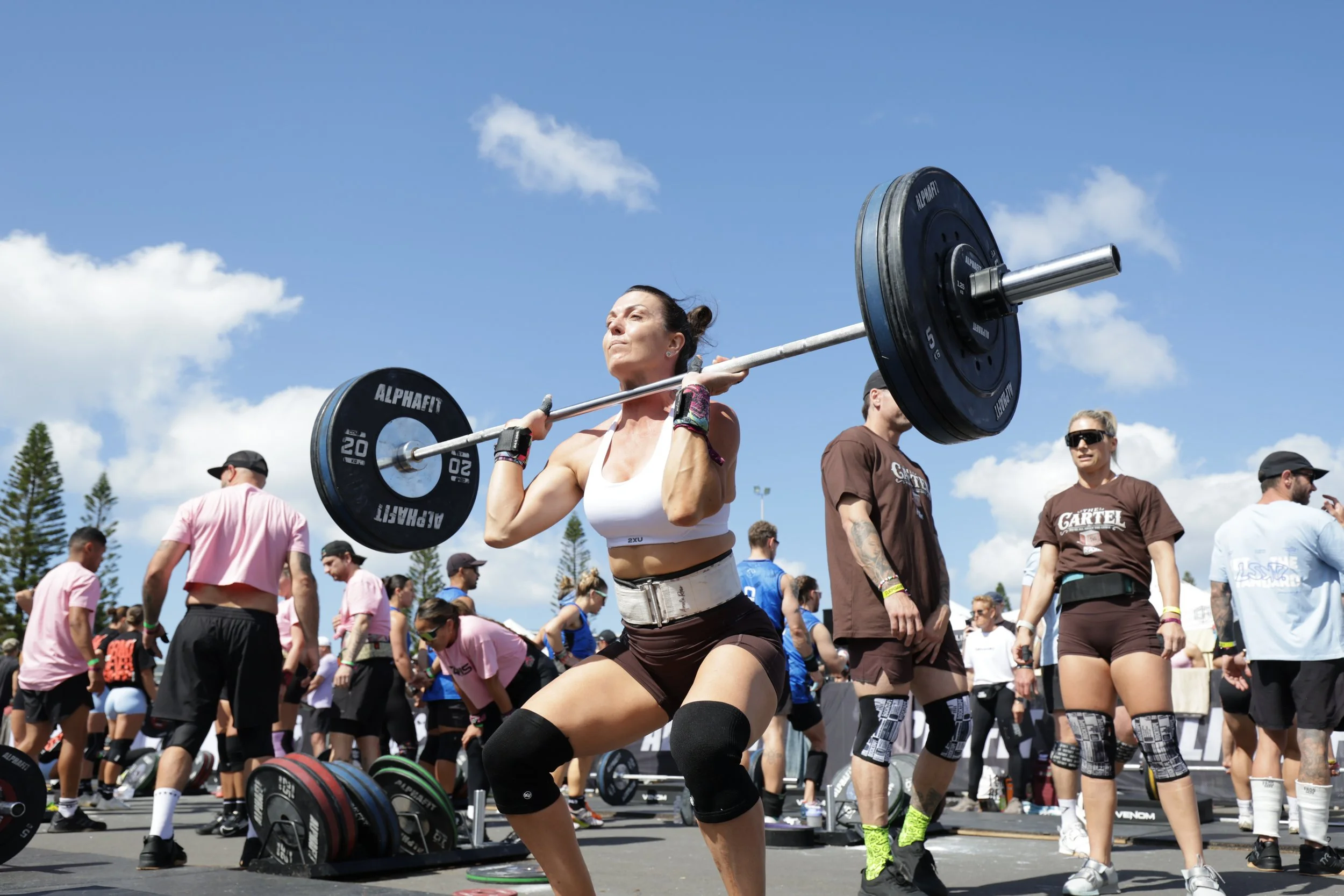 Woman lifting a barbell during a weightlifting competition outdoors with a crowd in the background.