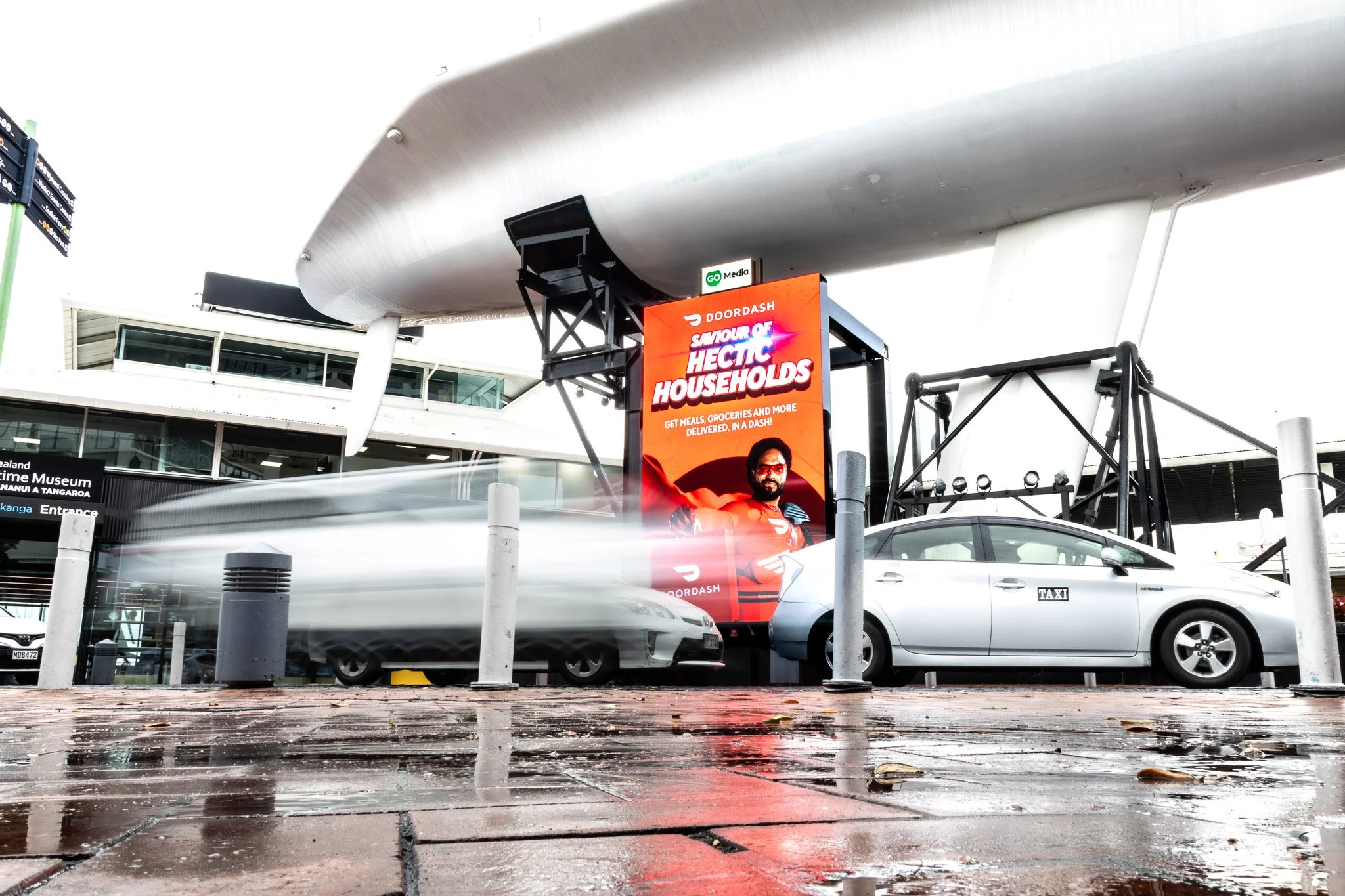 Outdoor urban scene with a large modern building, advertising billboard for DoorDash promoting 'Savour of Hectic Households', cars parked along the street, and wet pavement with fallen leaves, possibly after rain.