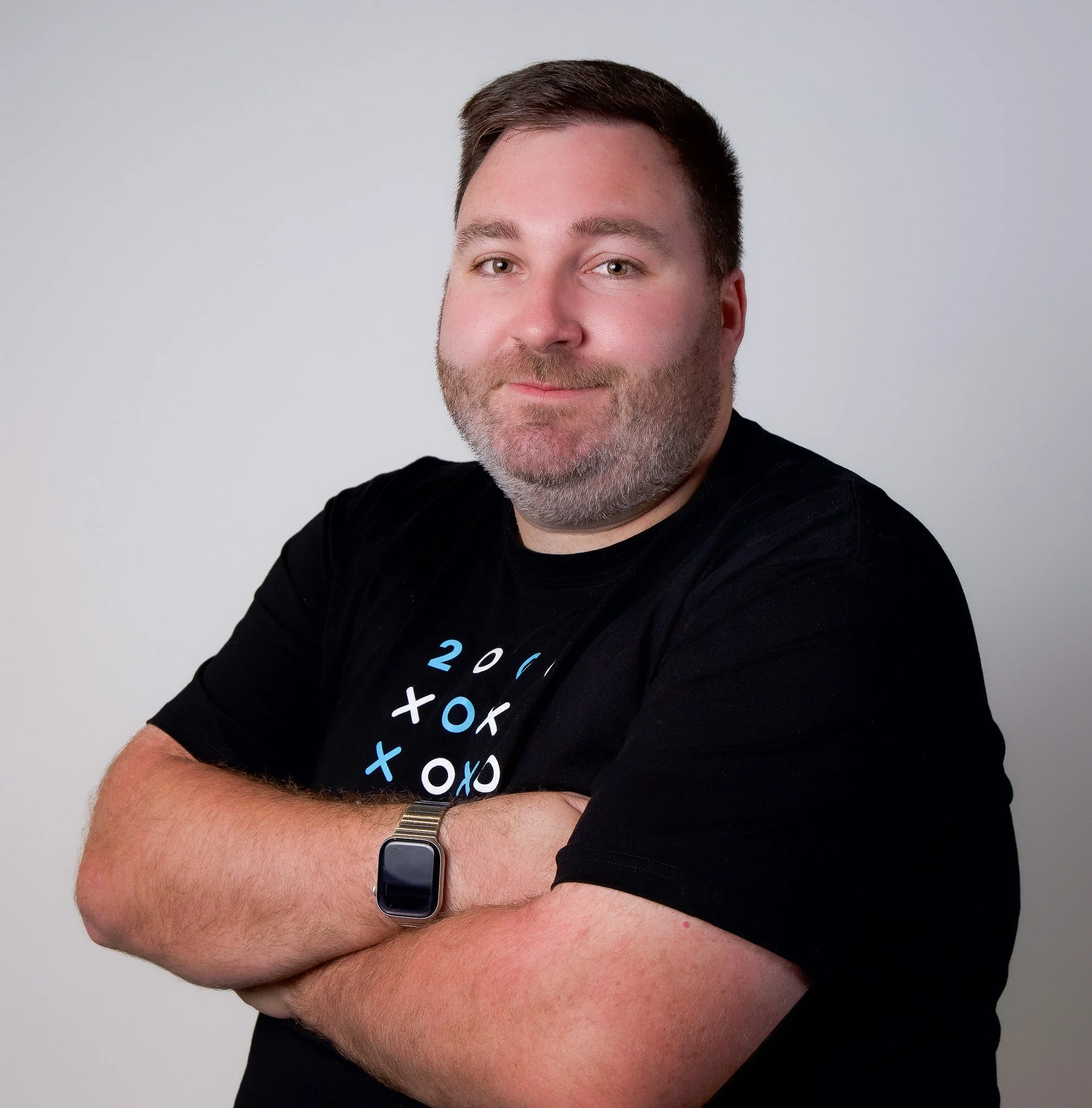 A man with short dark hair, a beard, and light skin, wearing a black T-shirt with colorful text, a wristwatch, and a smartwatch, stands with his arms crossed against a plain light gray background.