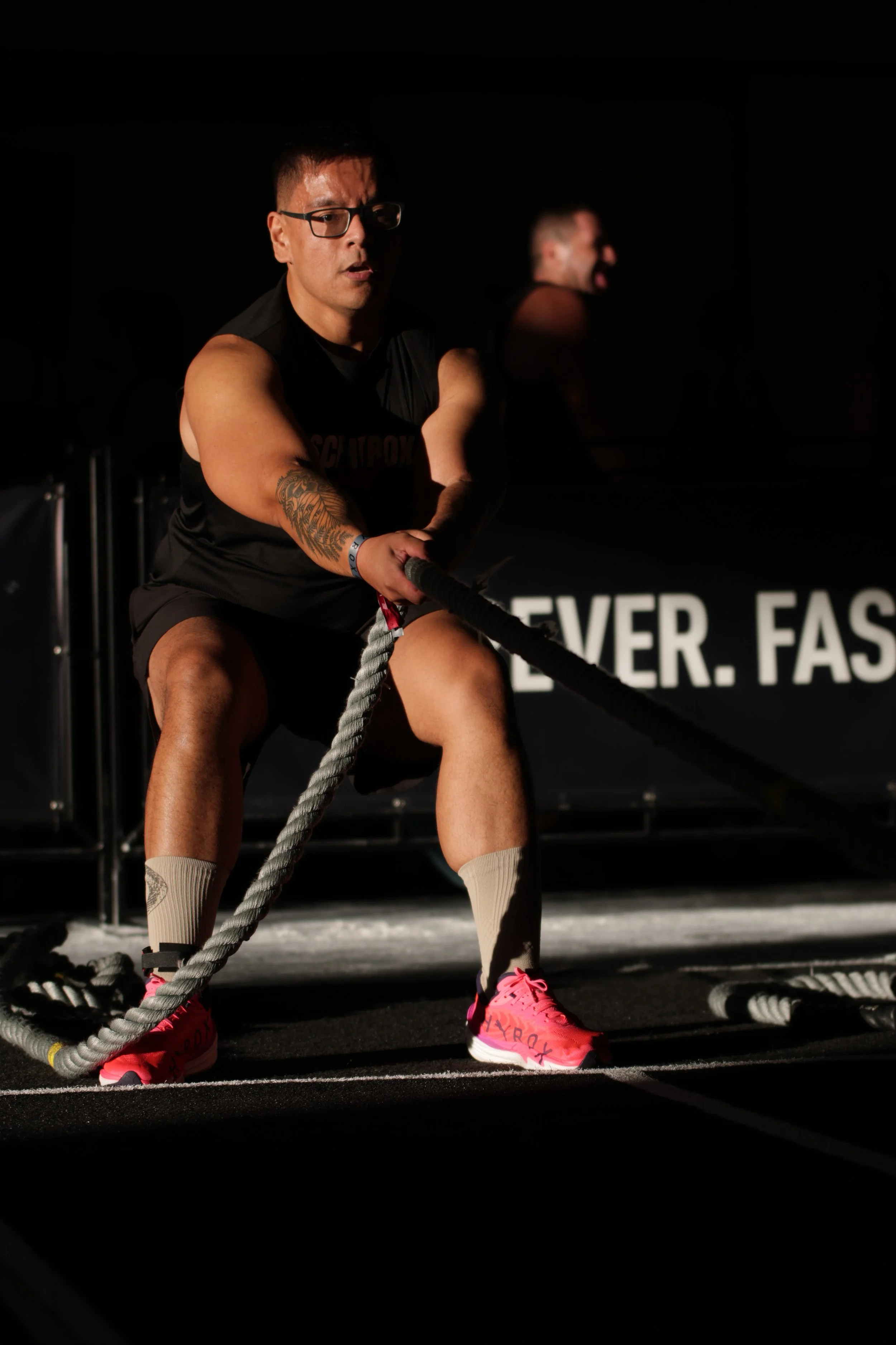 A man wearing glasses and a sleeveless shirt is using battle ropes in a gym. There is another man in the background. The gym has a black wall with white text, and the surroundings are dark.