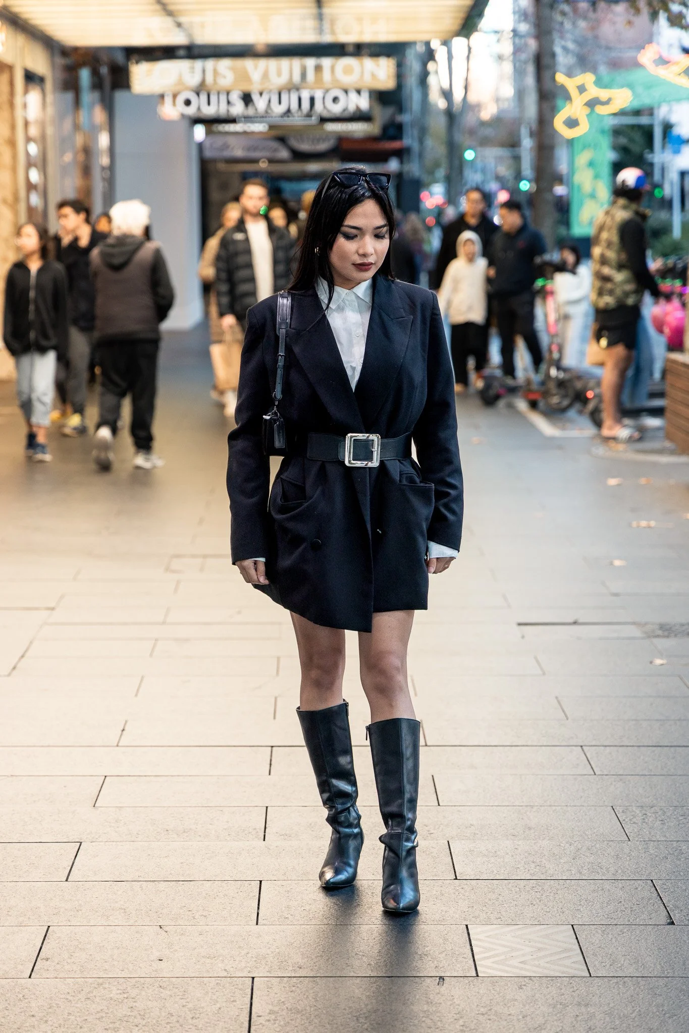 A woman walking in an urban shopping district at sunset, wearing a black blazer, white shirt, black knee-high boots, and sunglasses on her head.