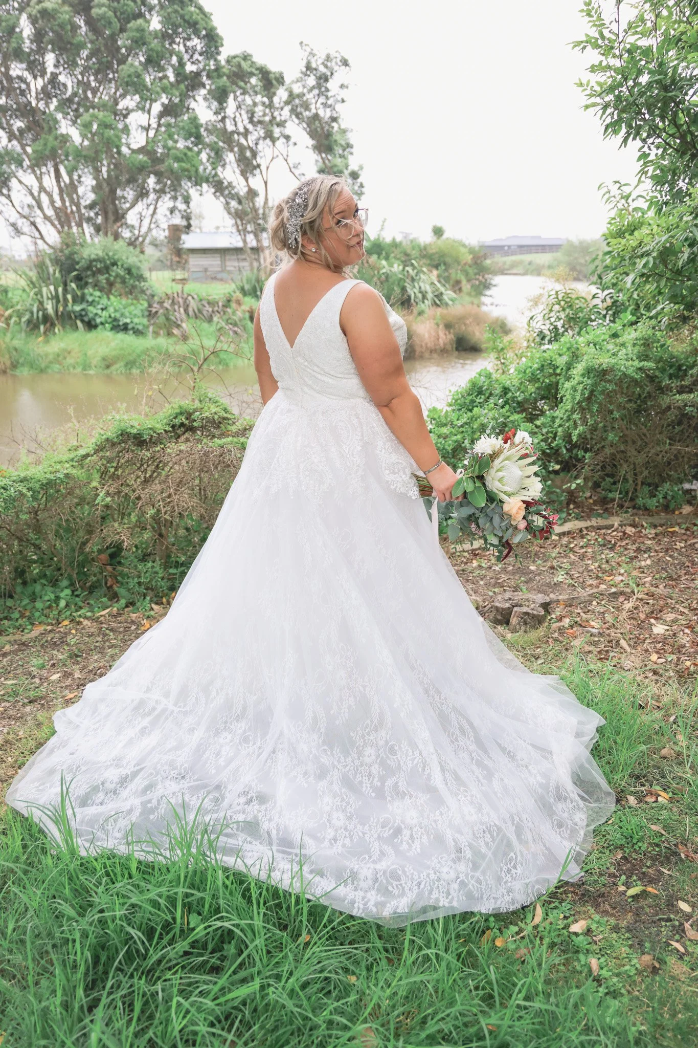 A bride standing outdoors near a pond, wearing a white lace wedding gown and holding a bouquet of flowers, with trees and a grassy area in the background.