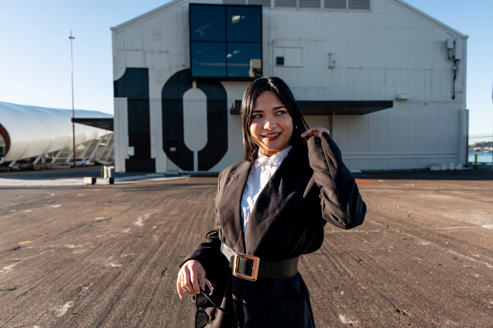 A woman in business attire standing outside on a tarmac in front of a large building with a black and white mural, with water and boats visible in the background.