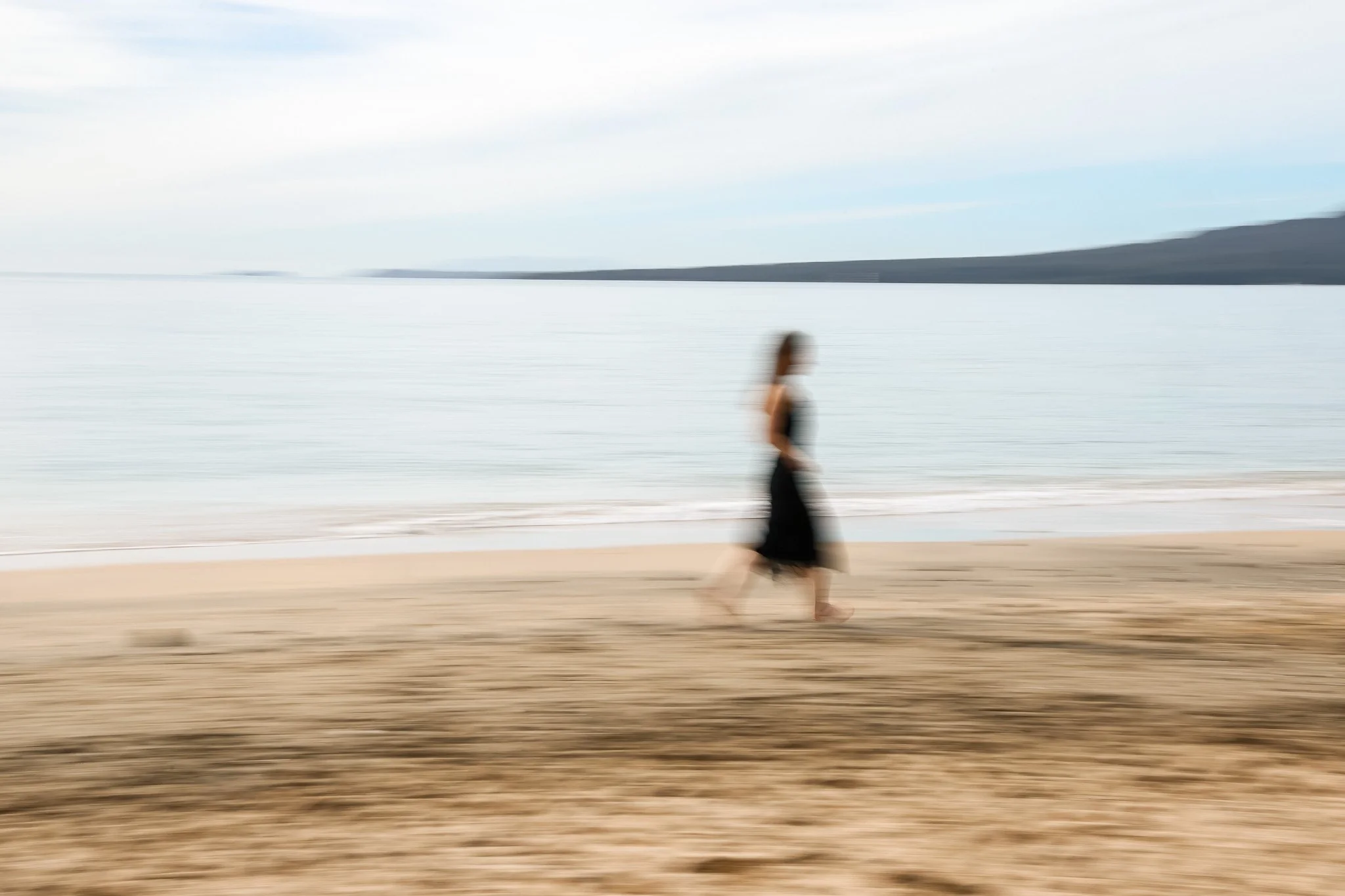A person walking on the beach with a blurred effect, with water and distant hills in the background.