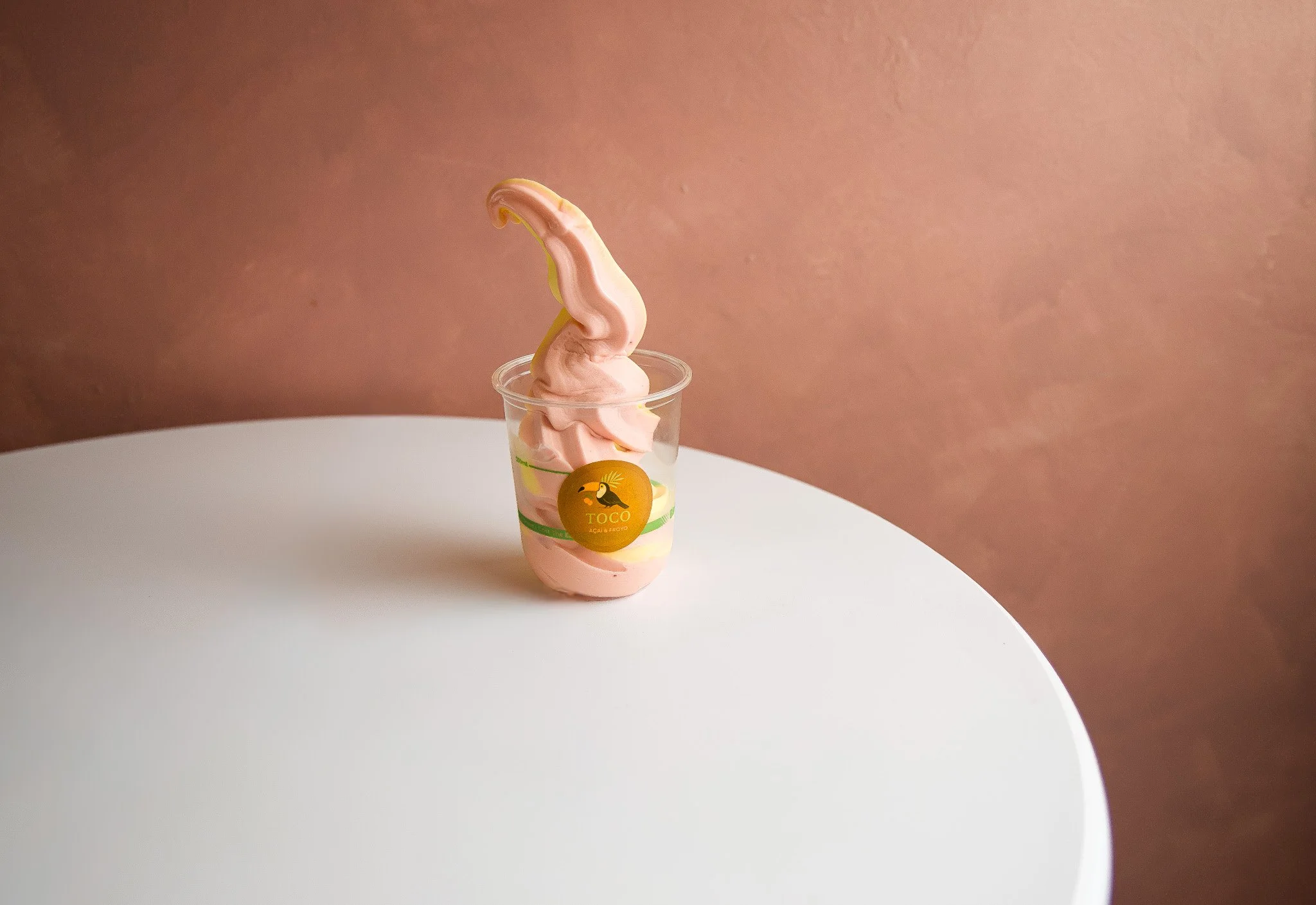 Soft serve ice cream in a clear cup with a green and yellow label, topped with a pink swirl and a decorative pink curl, on a white surface with a light pink wall background.