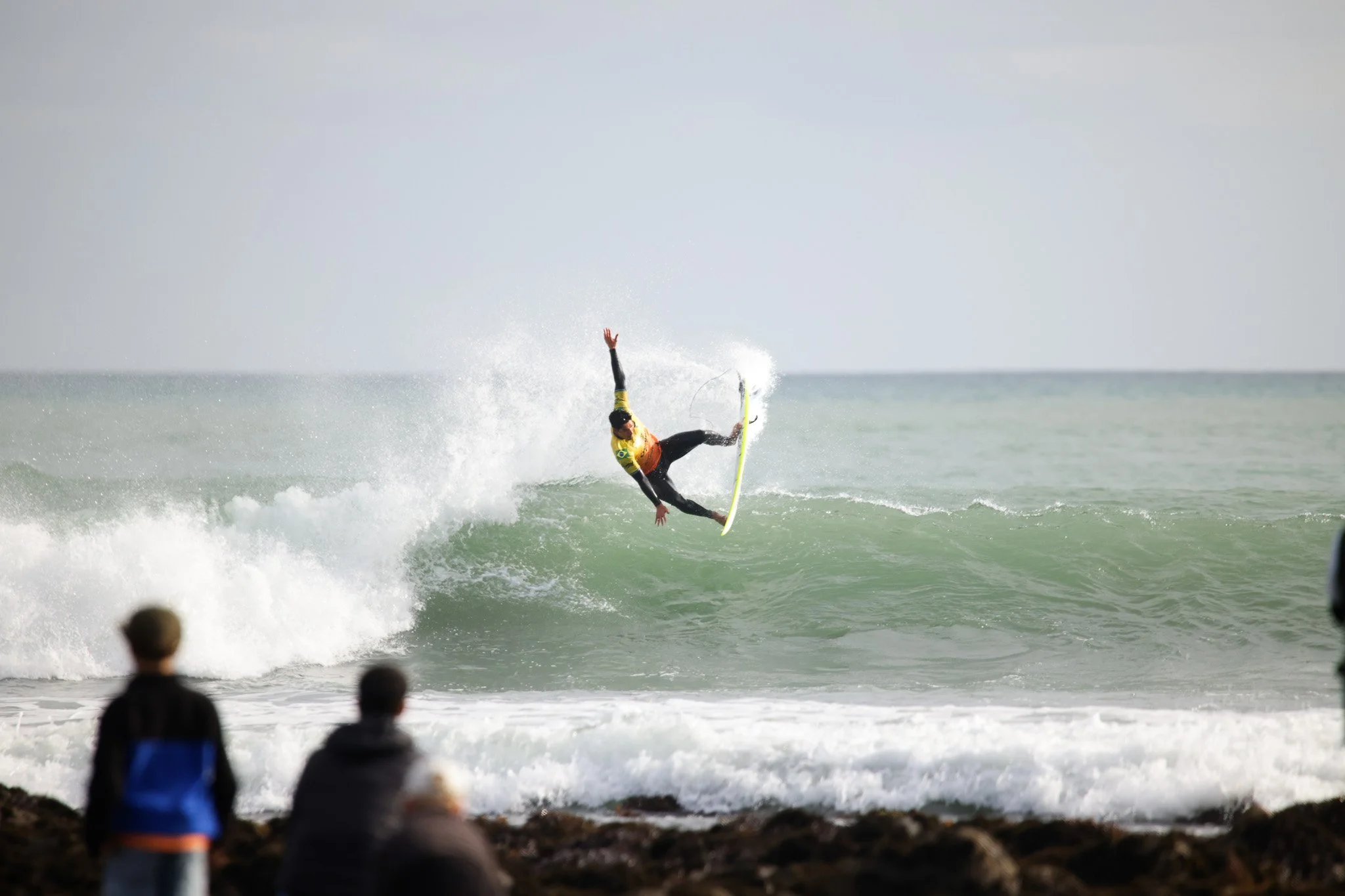 A surfer in a yellow shirt performing a trick on a wave at the beach with spectators watching from the shore.