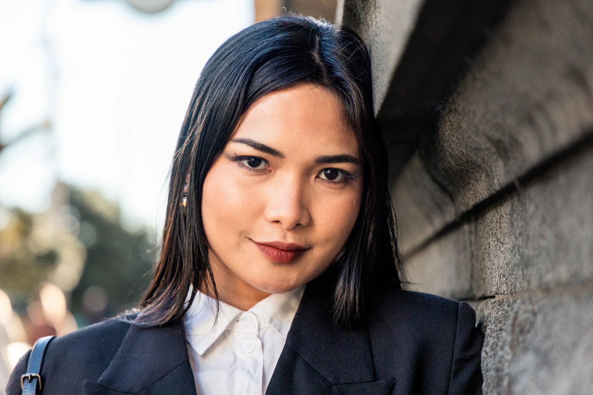 Close-up of a young woman with black hair, fair skin, wearing a dark blazer, white shirt, and a backpack, leaning against a brick wall outdoors.