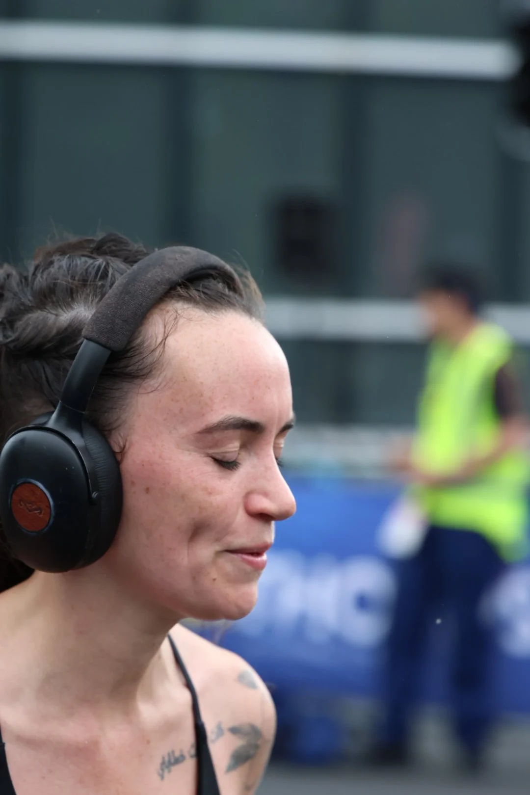 Close-up of a woman with curly brown hair, wearing large black headphones, with her eyes closed and a slight smile, outdoors with a blurred background and a person in a yellow safety vest.