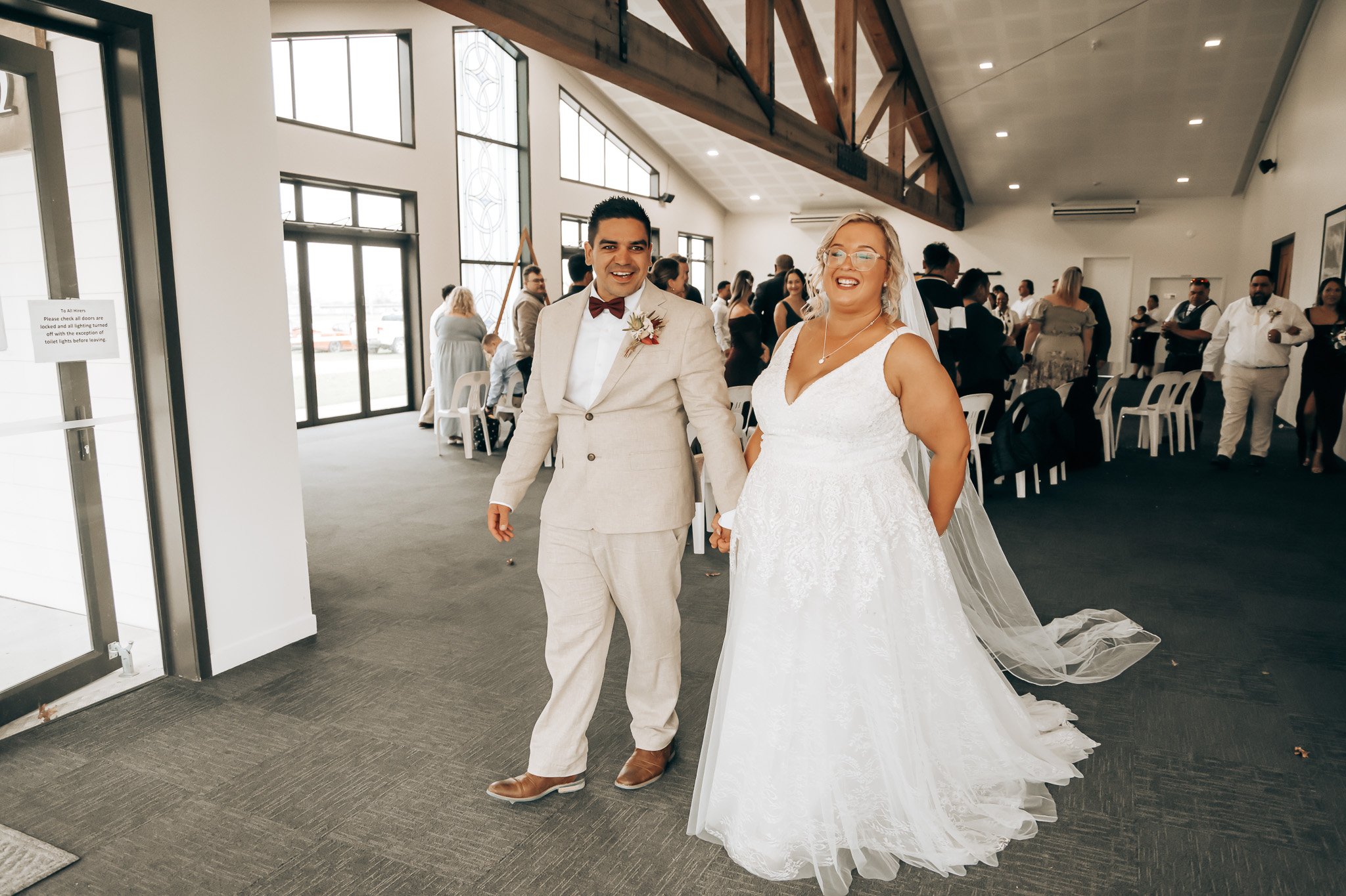 A newlywed couple holding hands and smiling inside a bright, modern venue with guests in the background. The groom is wearing a light-colored suit with a red bow tie and boutonniere, and the bride is in a white lace wedding gown with a long train and