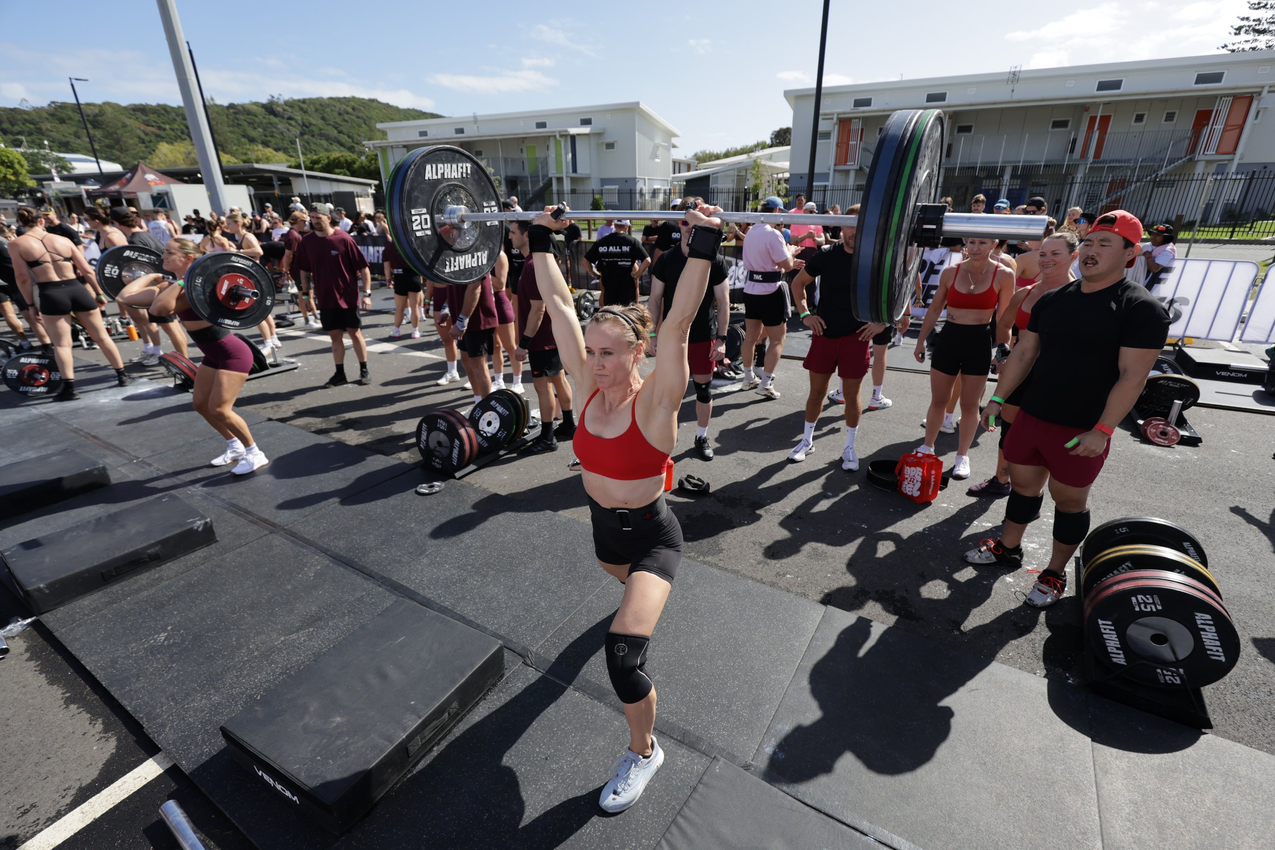 A woman in a red sports bra and black shorts lifts a barbell with weights during an outdoor fitness event, with numerous other participants and spectators around.