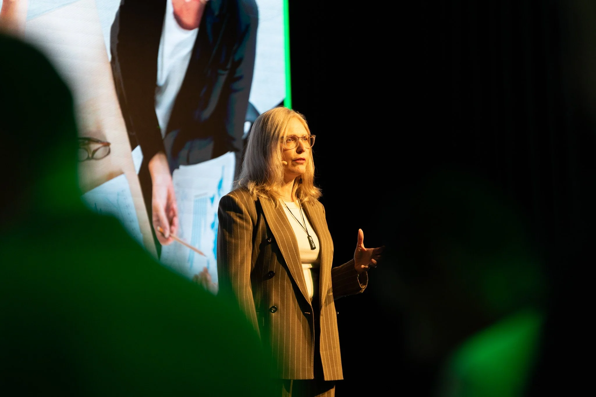 A woman with long blond hair, glasses, and a striped brown blazer speaking on stage, with a large screen behind her displaying a blurred image of a person.