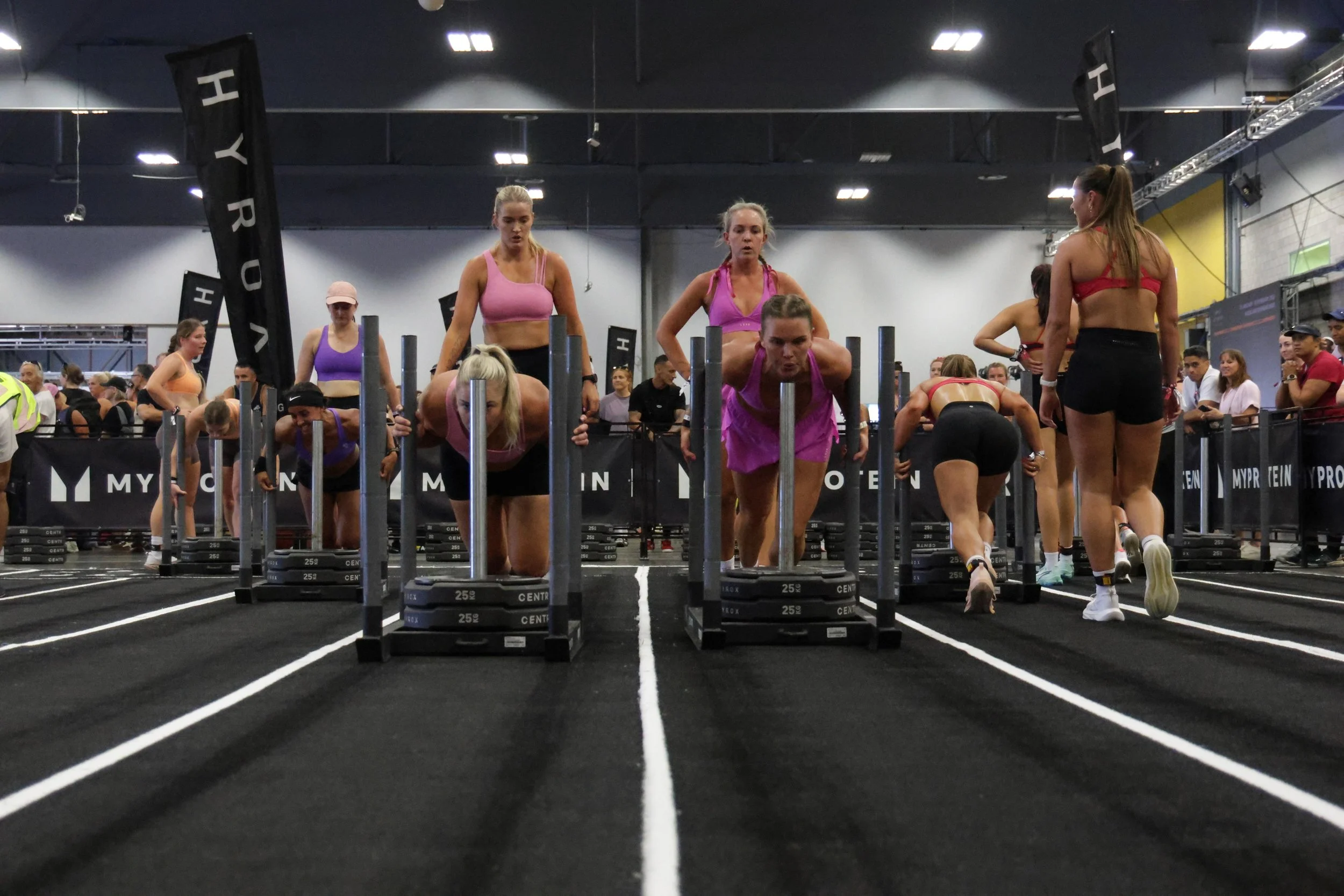 Women participating in a CrossFit competition, performing push-ups on a black turf with white lines, surrounded by spectators and event banners.