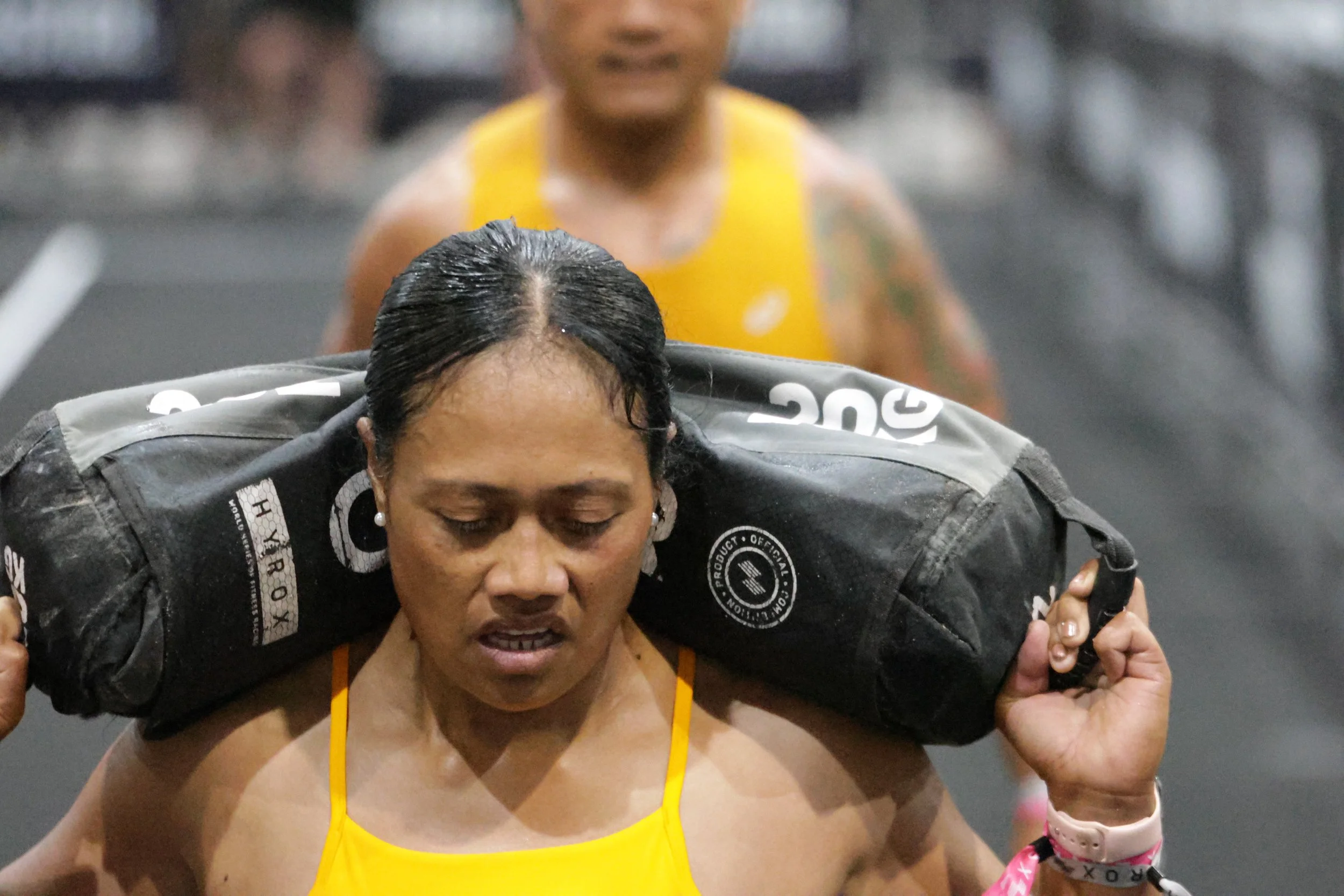 Two women pushing each other on a treadmill, likely during a workout or race, with a blurred background. One woman is in a yellow tank top, and the other in black top and a pink wristband.