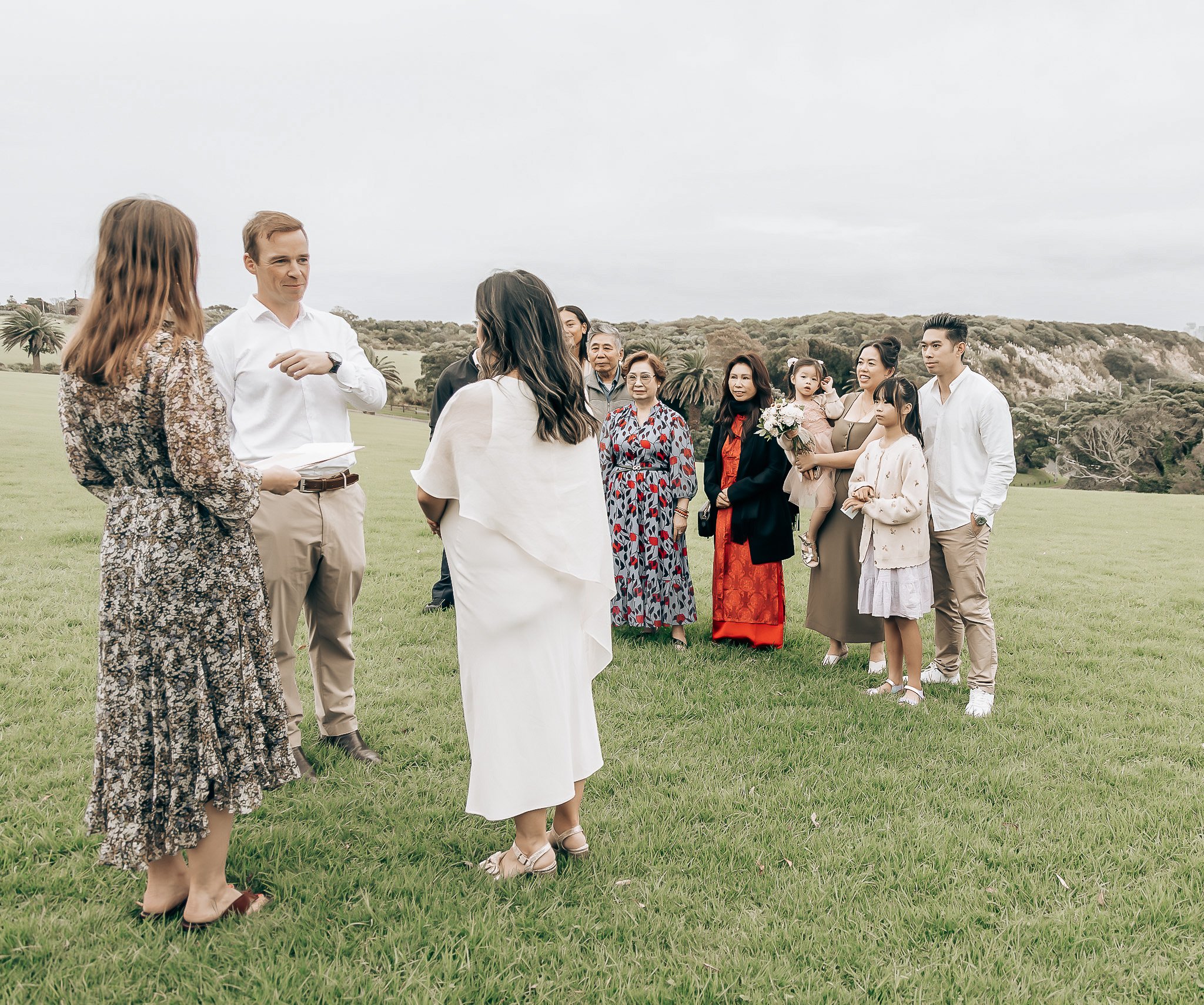 Group of people gathered outside on a cloudy day, with some individuals dressed in formal and traditional clothing, standing on a grassy field and engaging in conversation.