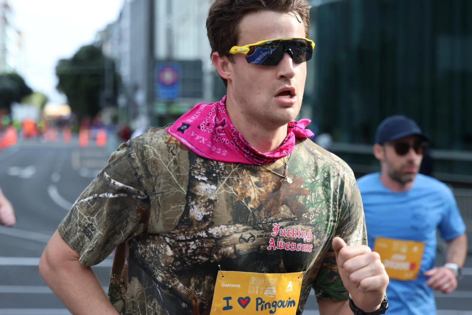 A young man running in a marathon, wearing sunglasses, a pink bandana, a camouflage t-shirt with the word "Fucking Awesome," and a yellow bib that says "I ❤️ Pingun," with other runners and orange traffic cones in the background.