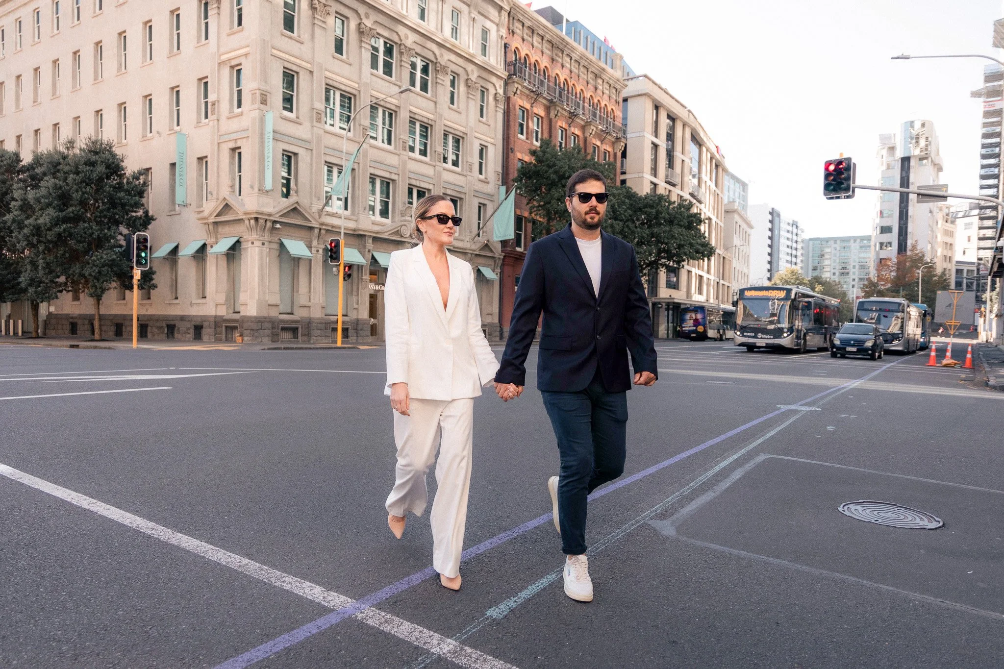 A man and woman walking hand in hand across a city street, both wearing sunglasses and dressed in formal attire, with traffic lights and buses in the background.