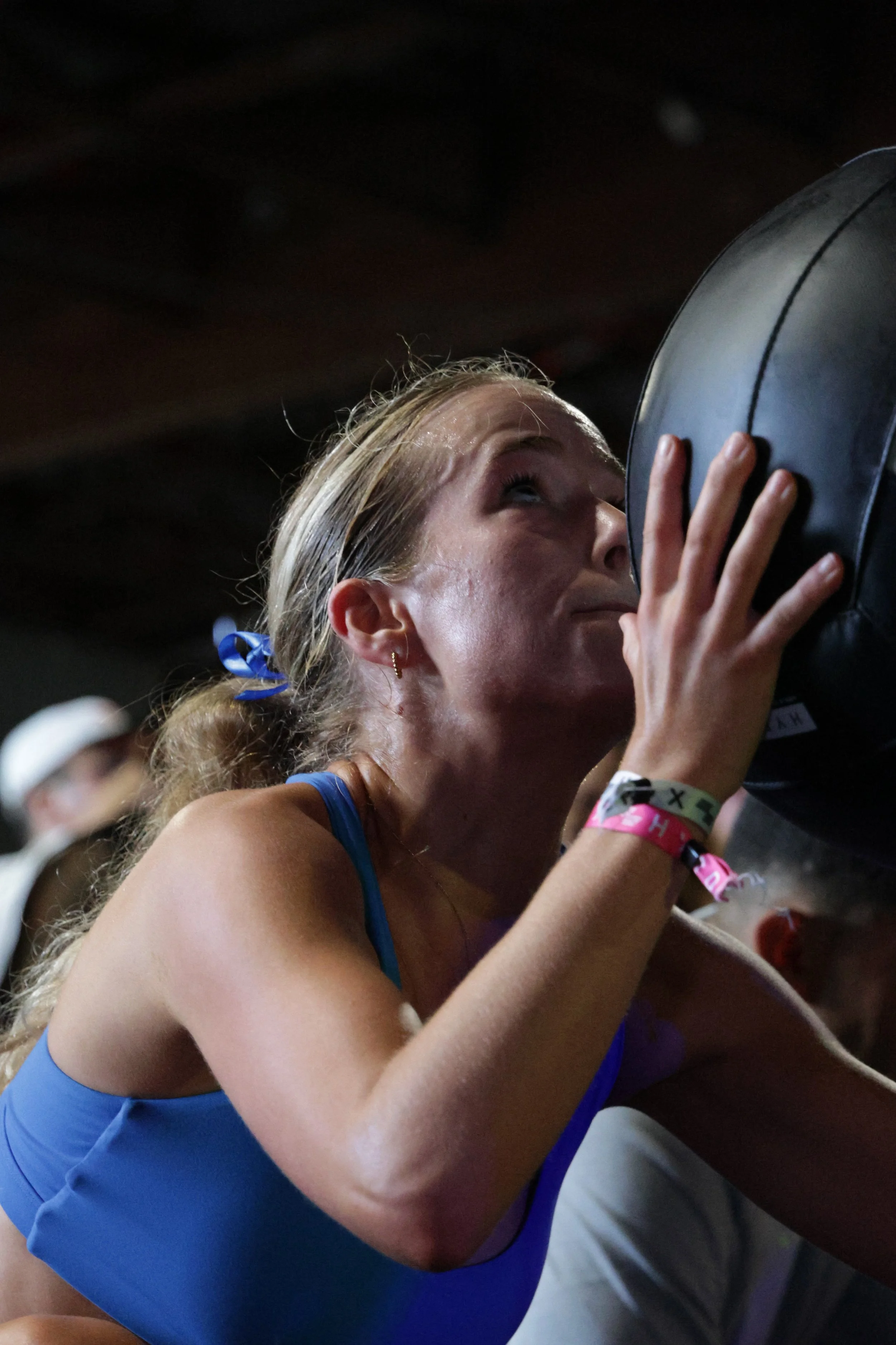 A female athlete in a blue sports bra, with her hair tied back, drinking water from a black water cooler or bottle, with a colorful wristband on her wrist, in a dimly lit setting.