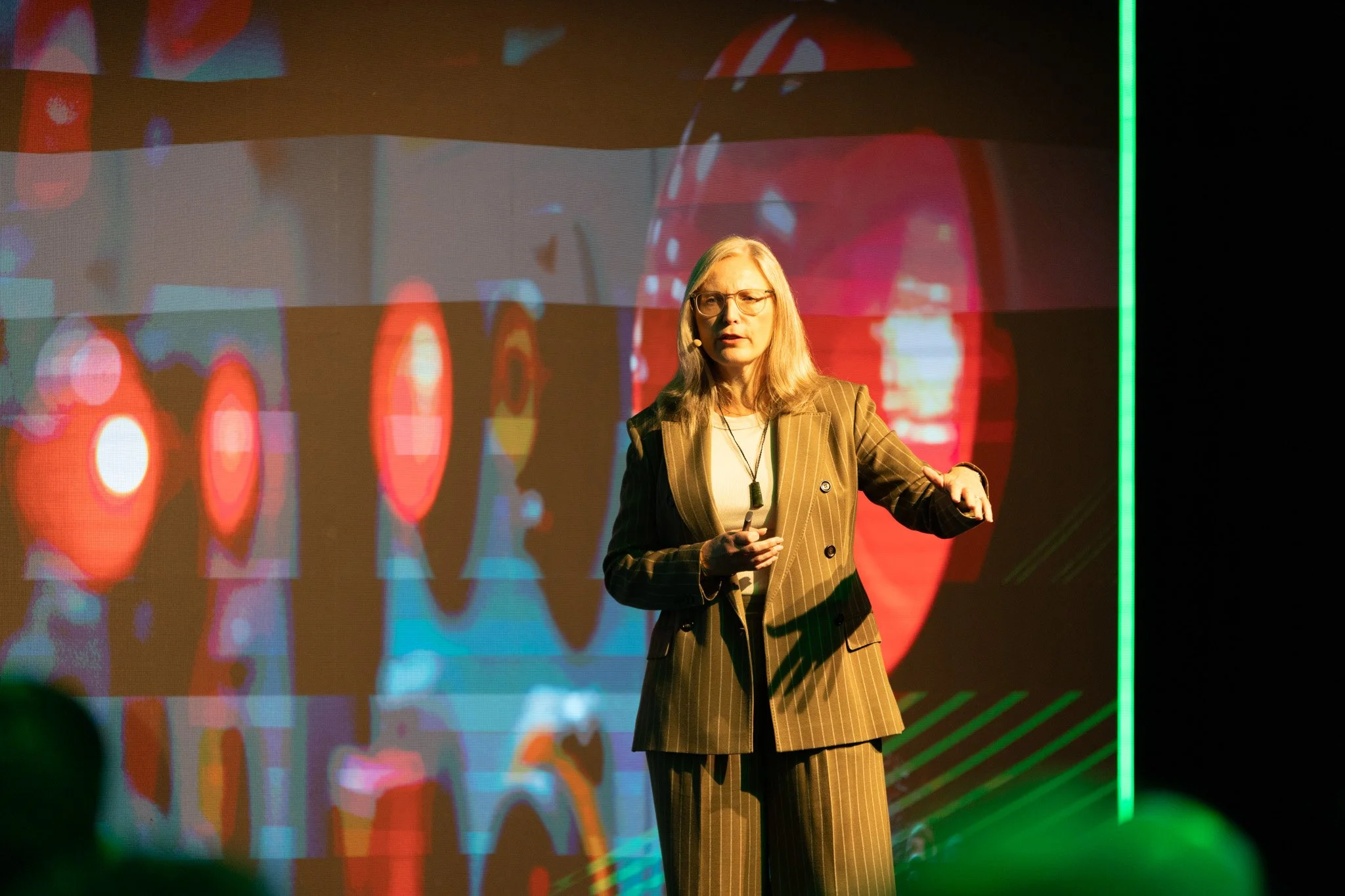 A woman with blonde hair and glasses giving a presentation on stage with a colorful abstract digital background displayed behind her.