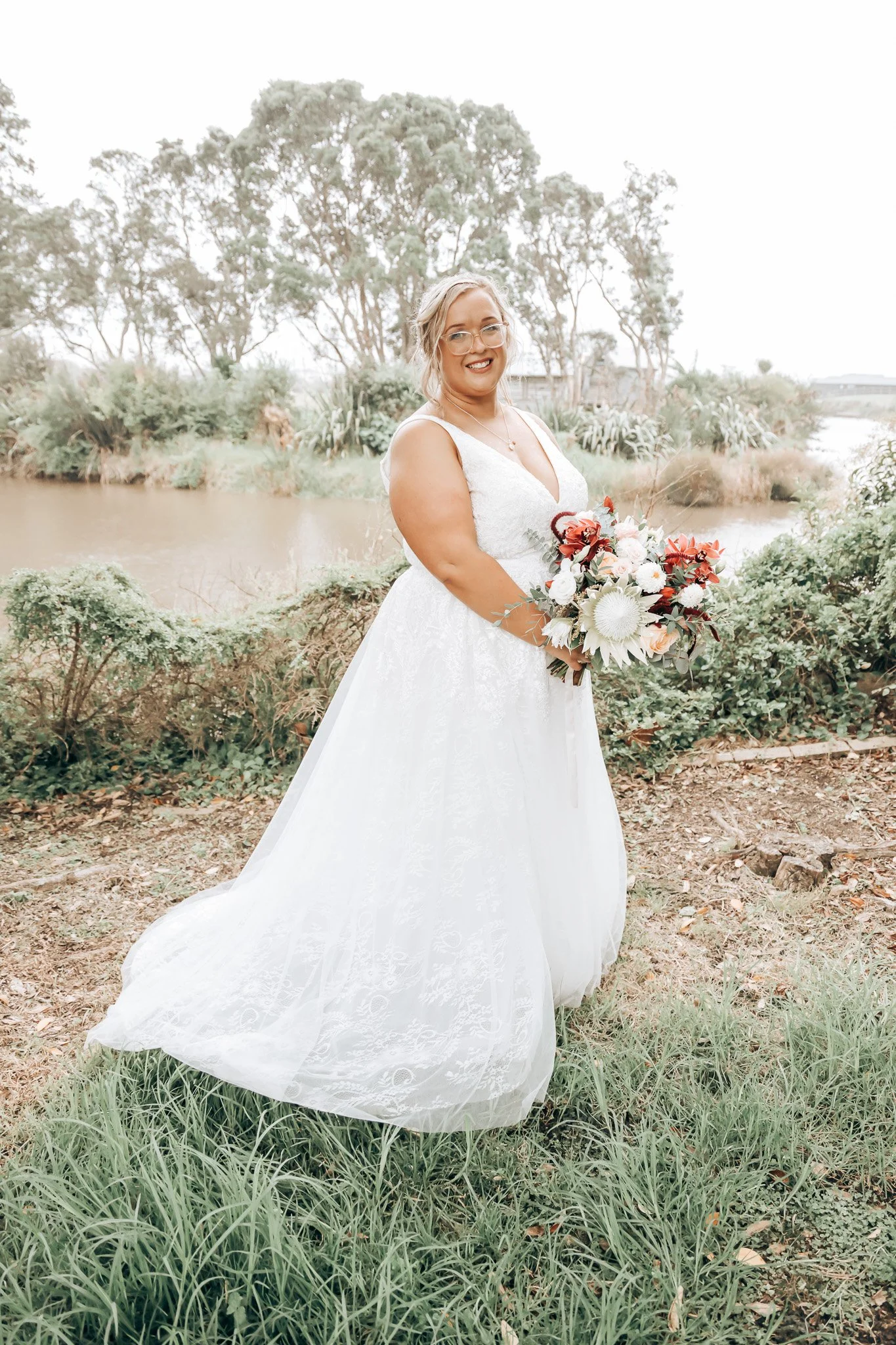 A woman in a white wedding dress holding a bouquet stands outdoors near a body of water, with trees and greenery in the background.