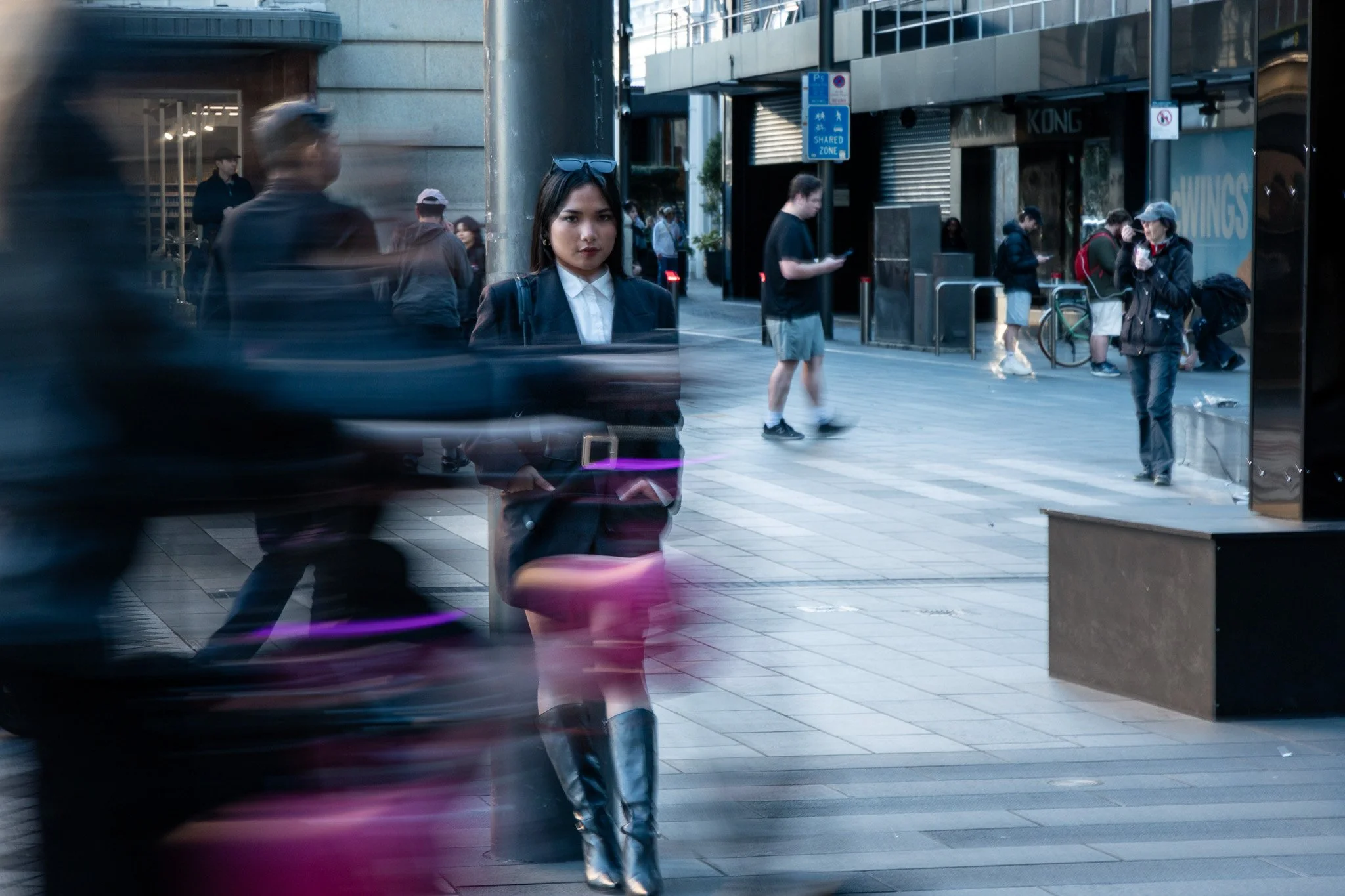 City street scene reflected in glass storefront showing pedestrians, including a woman in a black blazer with sunglasses on her head, walking and standing amid others on the sidewalk.