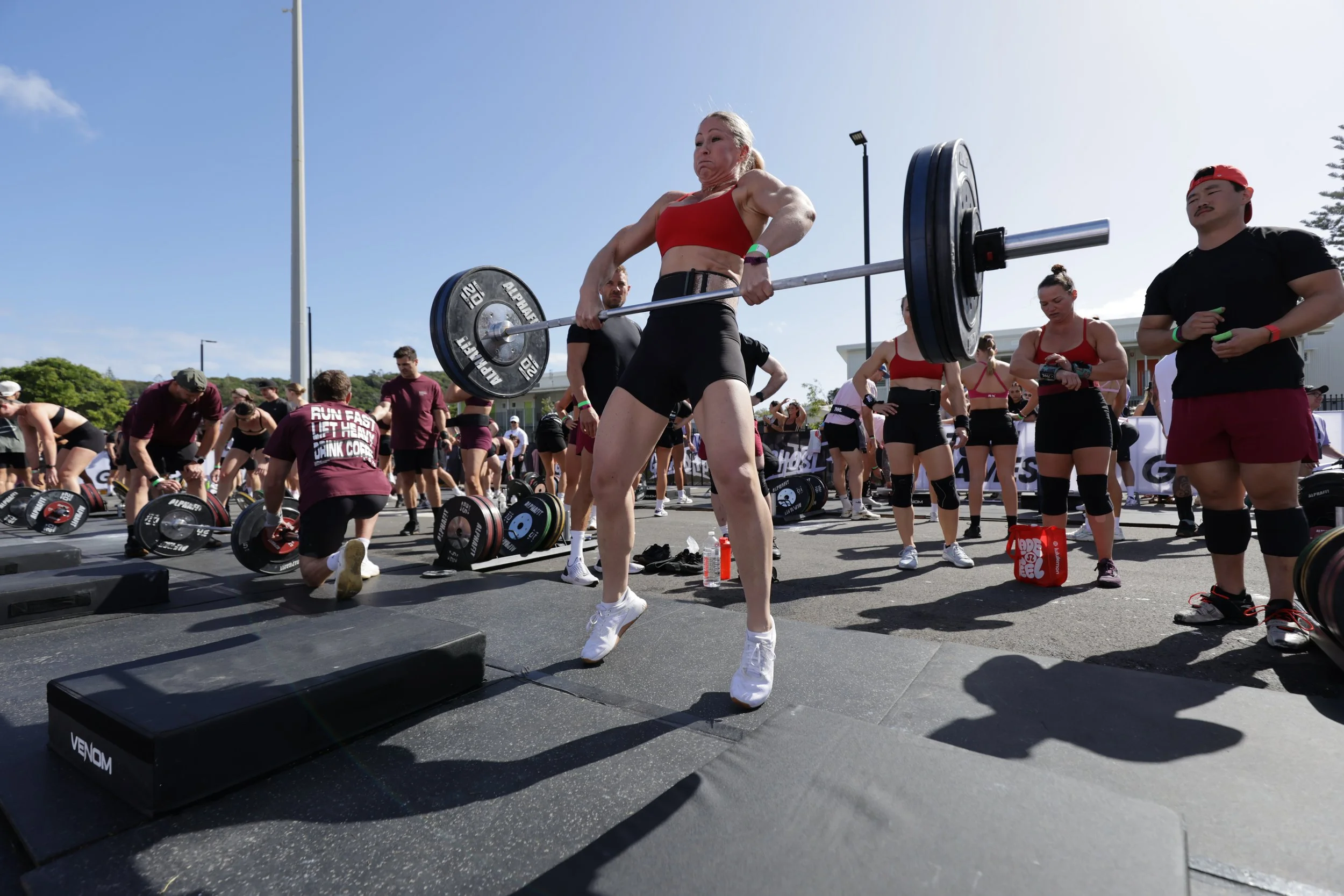 A woman in a red sports bra and black shorts is lifting a barbell with weights during an outdoor fitness event, surrounded by other participants and spectators.