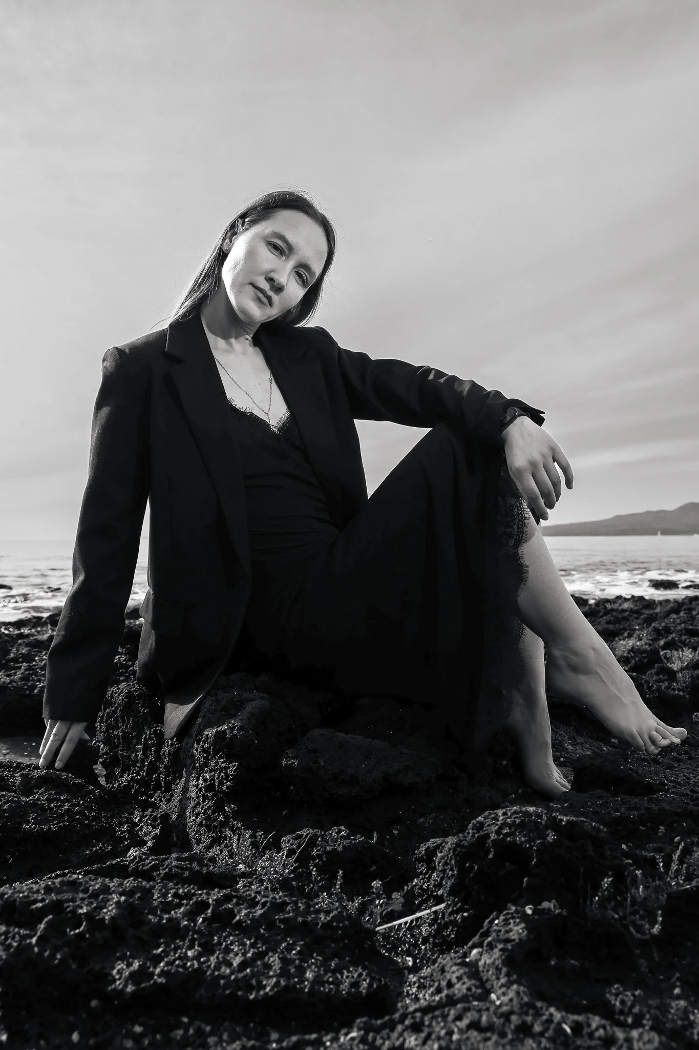A woman with long hair, wearing a blazer and lace dress, sitting on rocks by the sea during sunset or sunrise, in black and white.