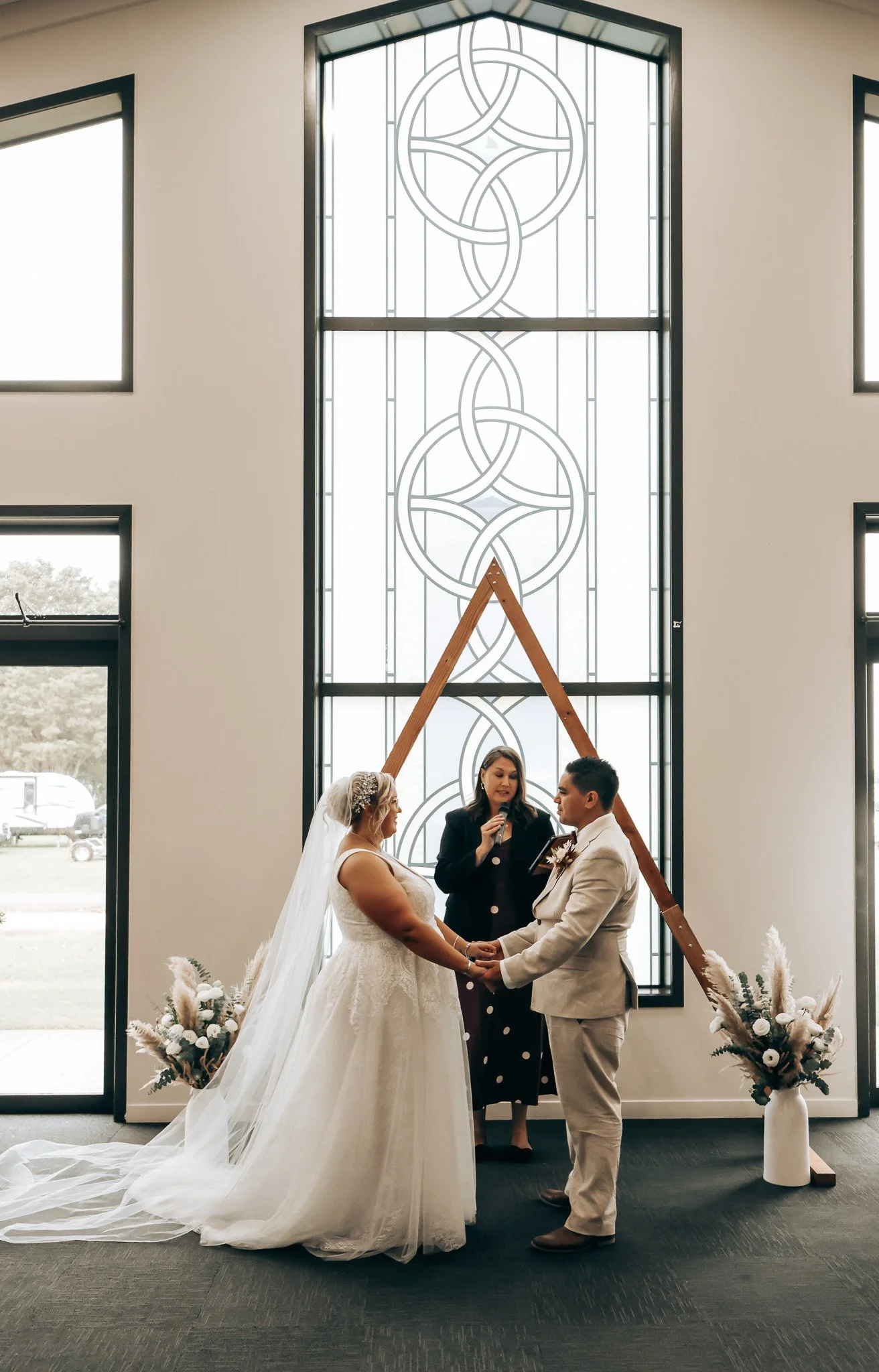 A wedding ceremony inside a church with a bride and groom holding hands, a minister officiating, and a large decorative stained glass window behind them. Flowers are arranged around the ceremony area.