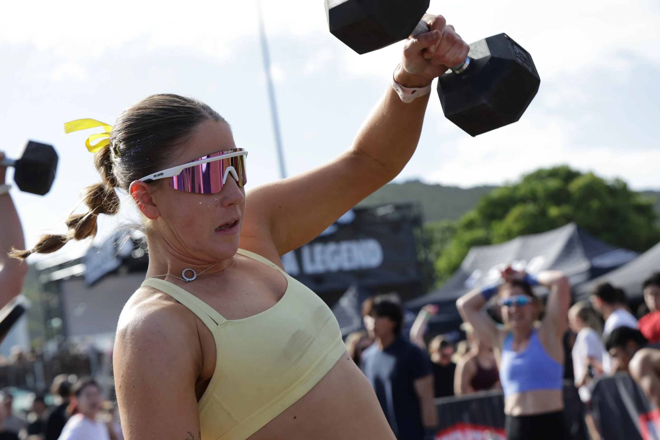 A woman wearing reflective sunglasses and a yellow sports bra lifting a dumbbell during an outdoor fitness event, with a crowd and tents in the background.