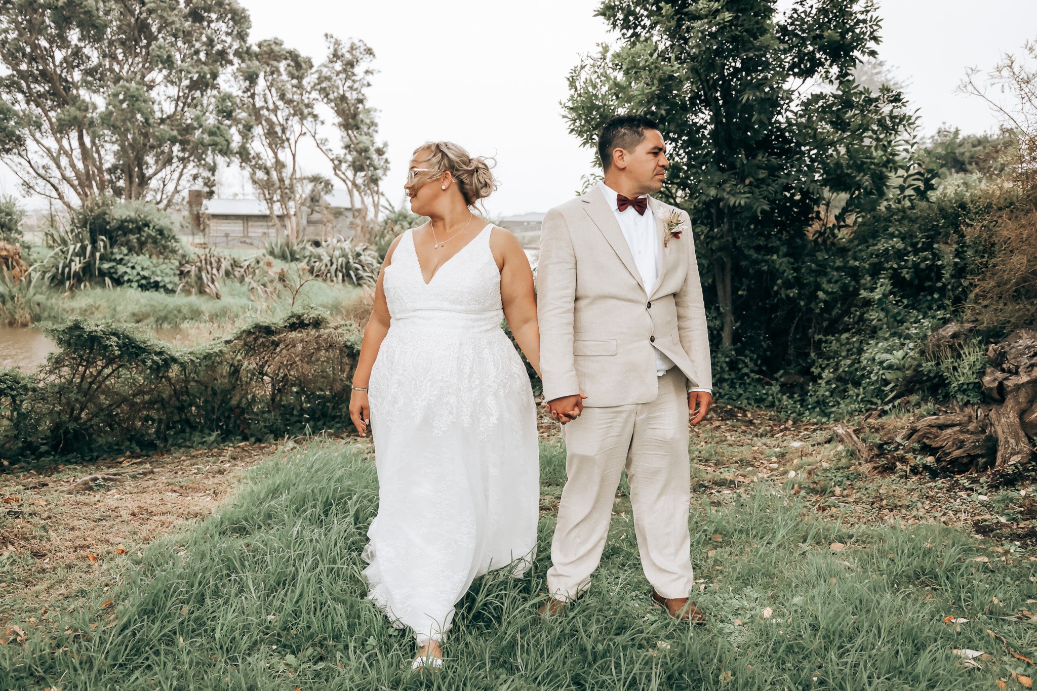 A bride and groom holding hands in an outdoor setting with trees and greenery in the background. The bride is wearing a white wedding dress with lace details, and the groom is dressed in a light beige suit with a white shirt and a burgundy bow tie.