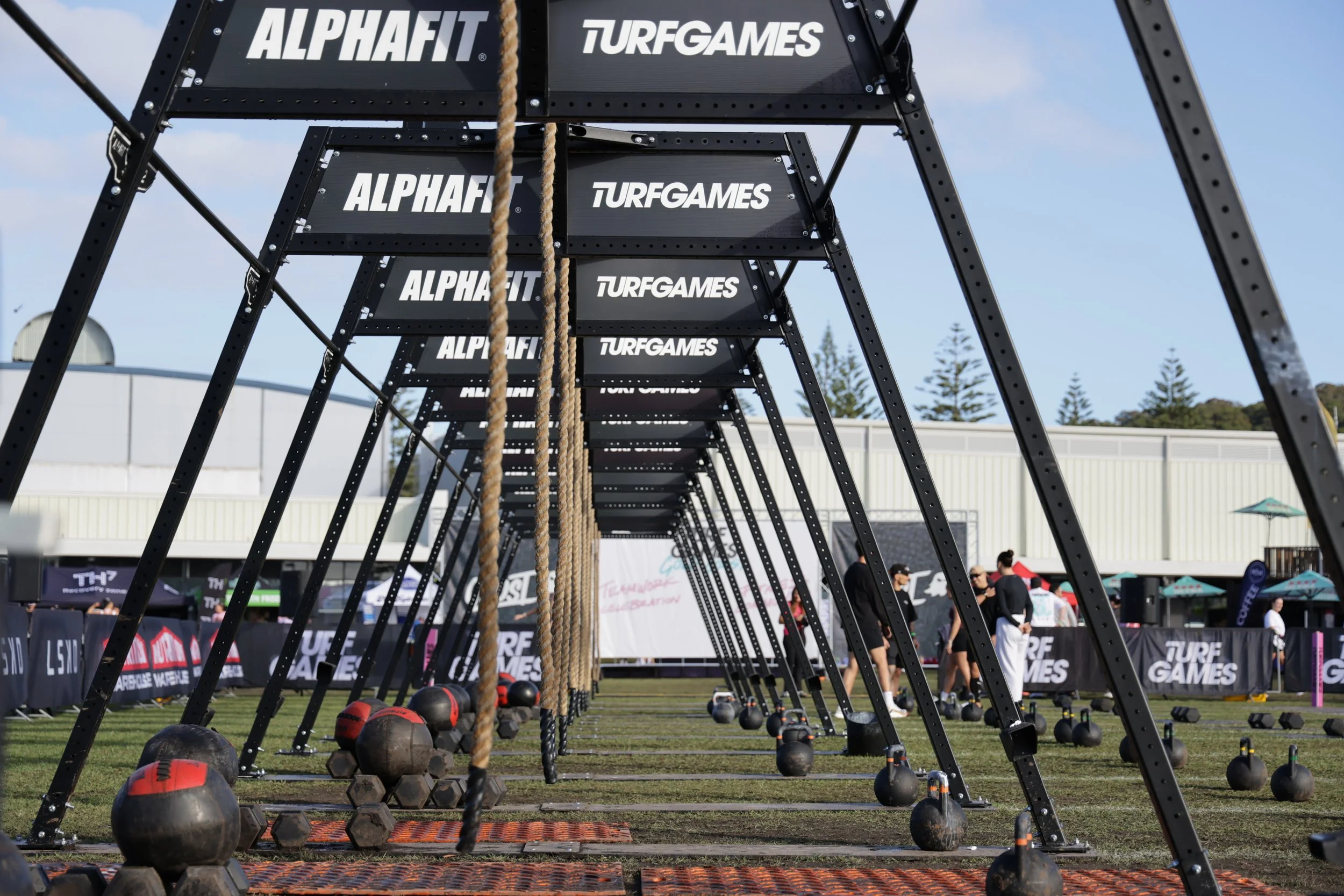 Outdoor fitness and obstacle course setup with black metal frames, ropes, and weighted balls, under a blue sky at a public event.