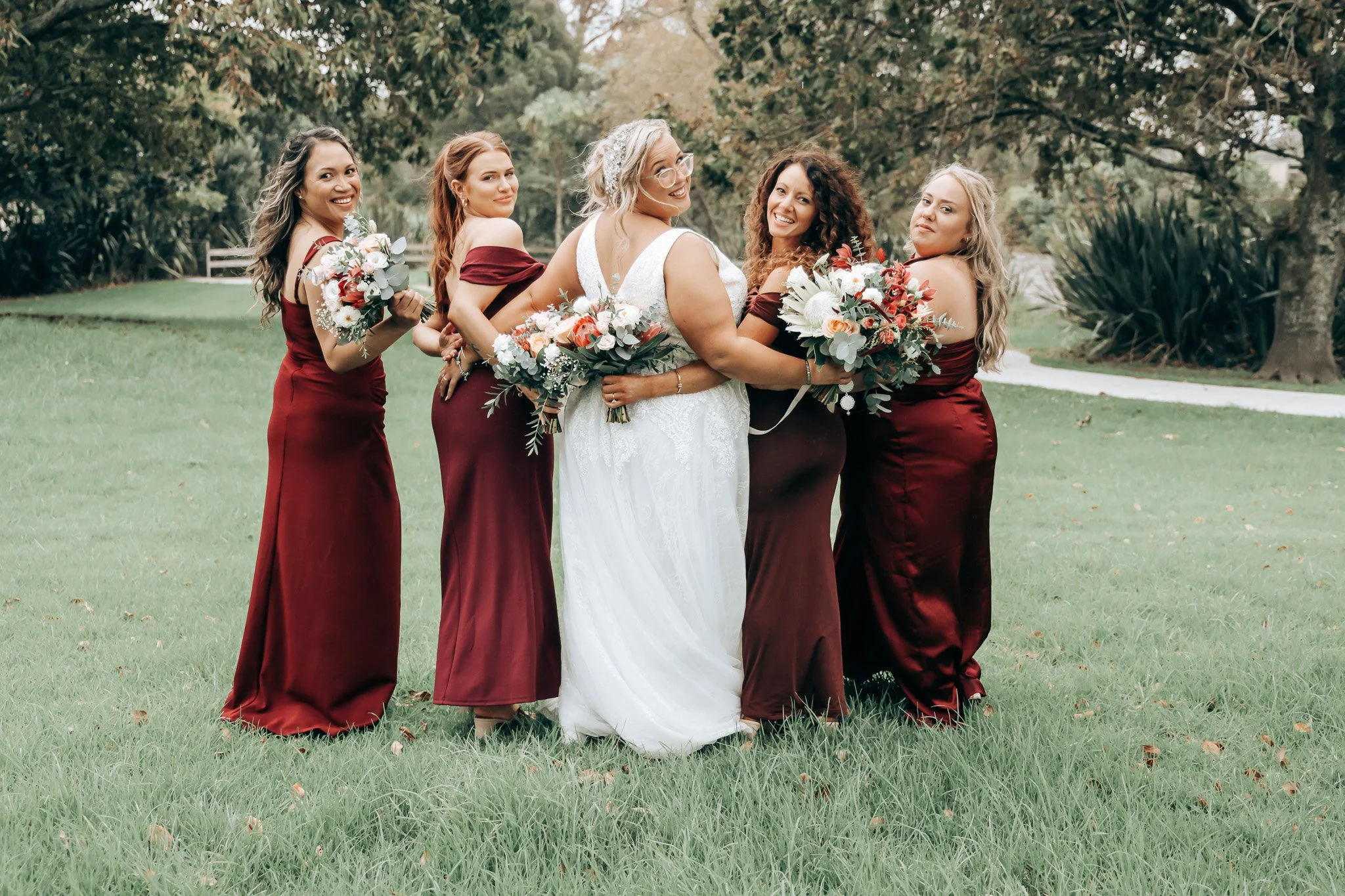 A bride in a white wedding dress is standing outdoors with five bridesmaids dressed in matching burgundy gowns, holding bouquets of flowers, with green grass and trees in the background.
