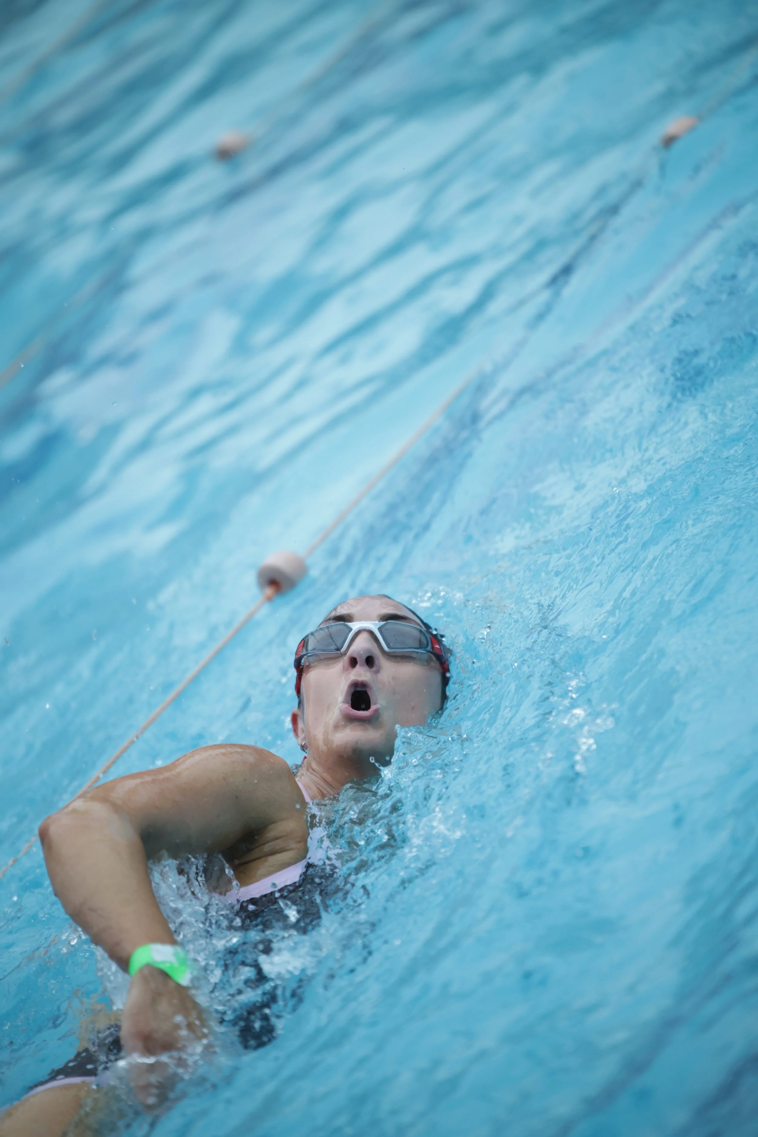 Swimmer in a pool, wearing goggles and a bathing suit, with an expression of effort or surprise, surrounded by blue water and lane dividers.