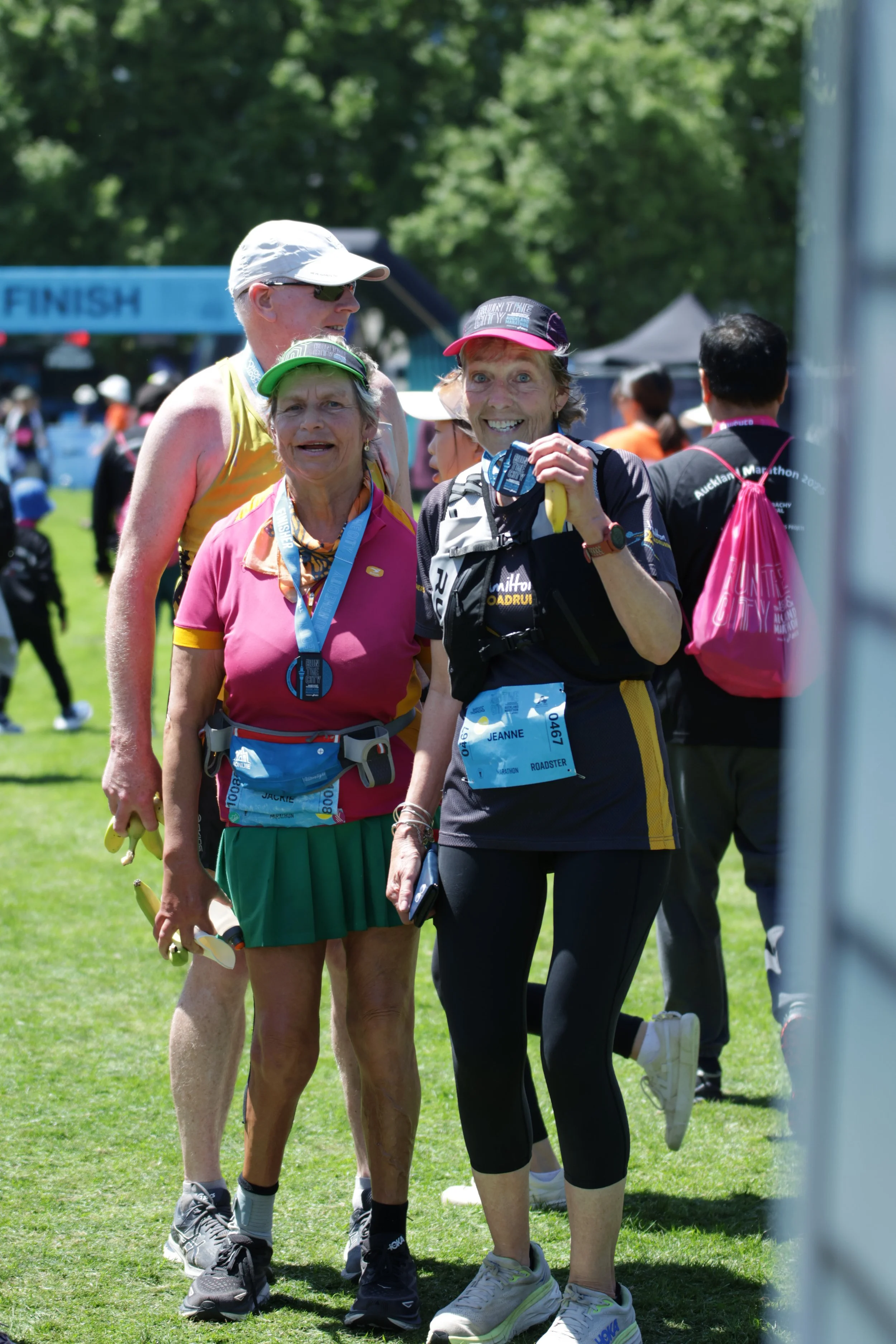 Three women standing together after a race, wearing medals and running gear, smiling with a finish line visible in the background on a sunny day.