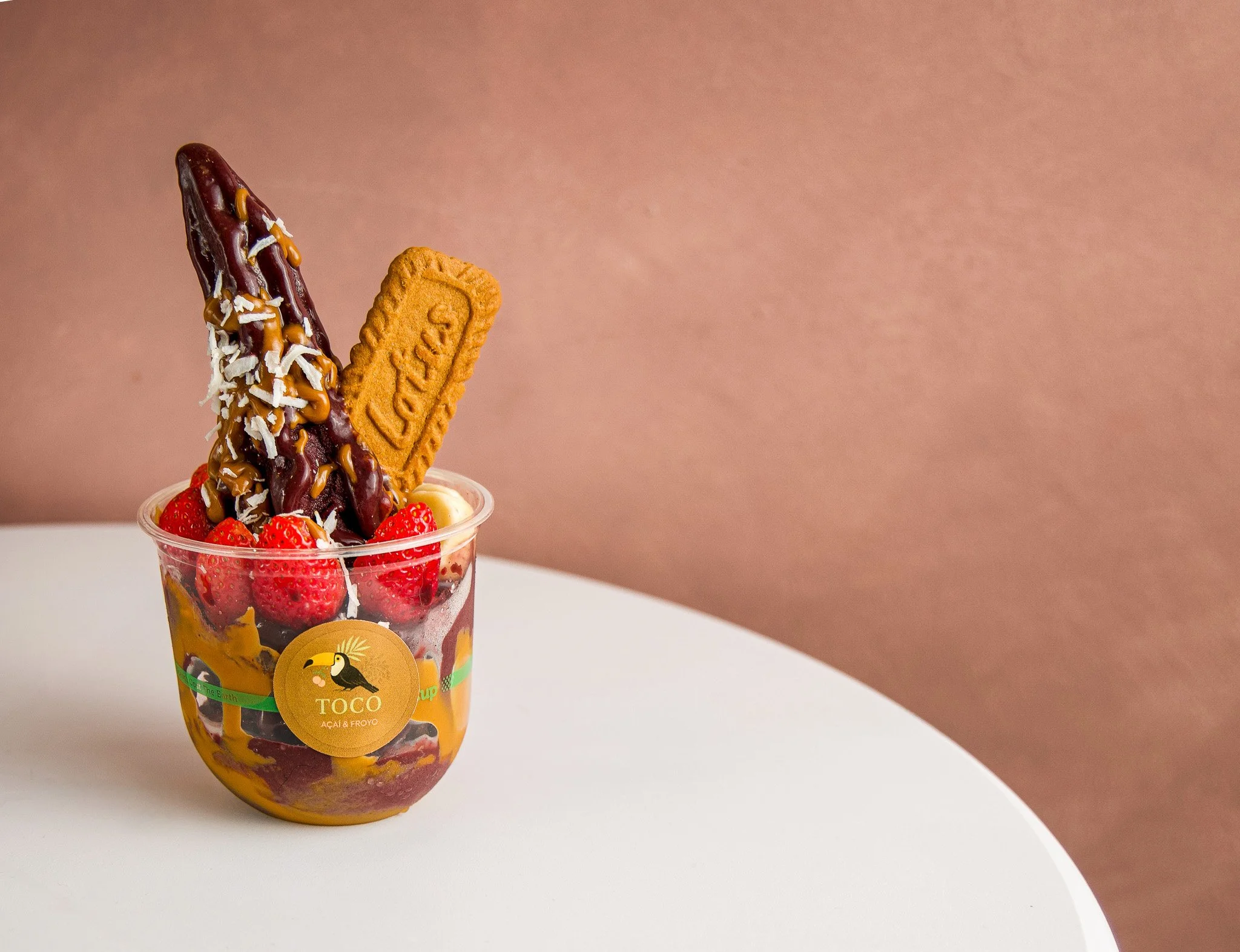 A clear plastic cup filled with strawberries, acai, froyo, and topped with a chocolate-covered banana, shredded coconut, and a biscuit.