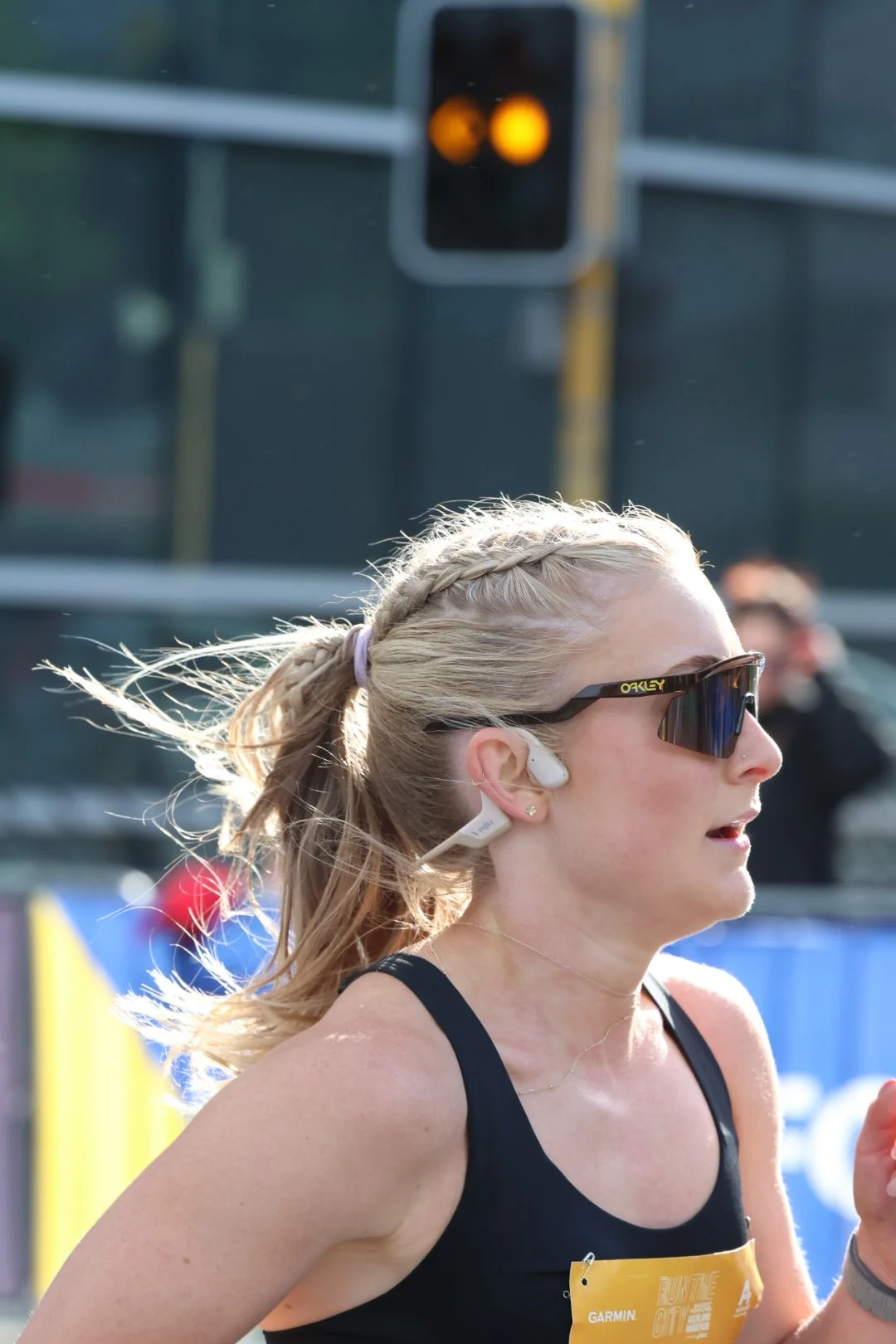 A female marathon runner wearing sunglasses and wireless earbuds, mid-race with wind in her hair, side profile, in an outdoor setting with a blurred background including a traffic light and onlookers.