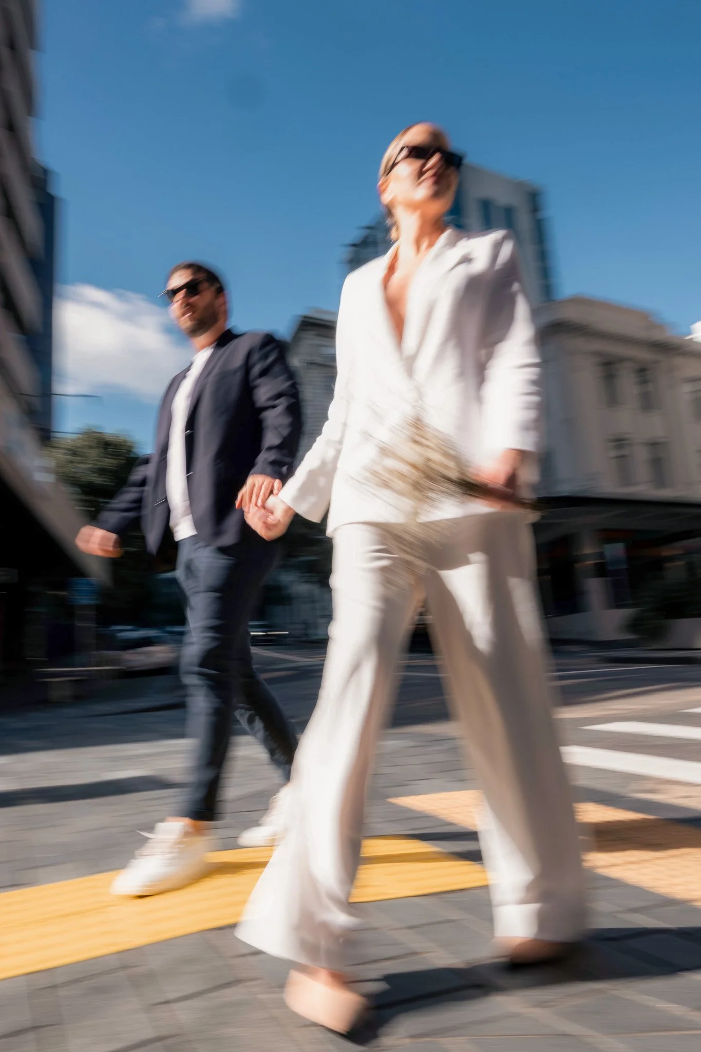 Blurred image of a woman in a white suit walking hand-in-hand with a man in a dark suit crossing the street at a pedestrian crosswalk in an urban environment. The woman is wearing sunglasses and the scene is set on a sunny day with buildings in the b