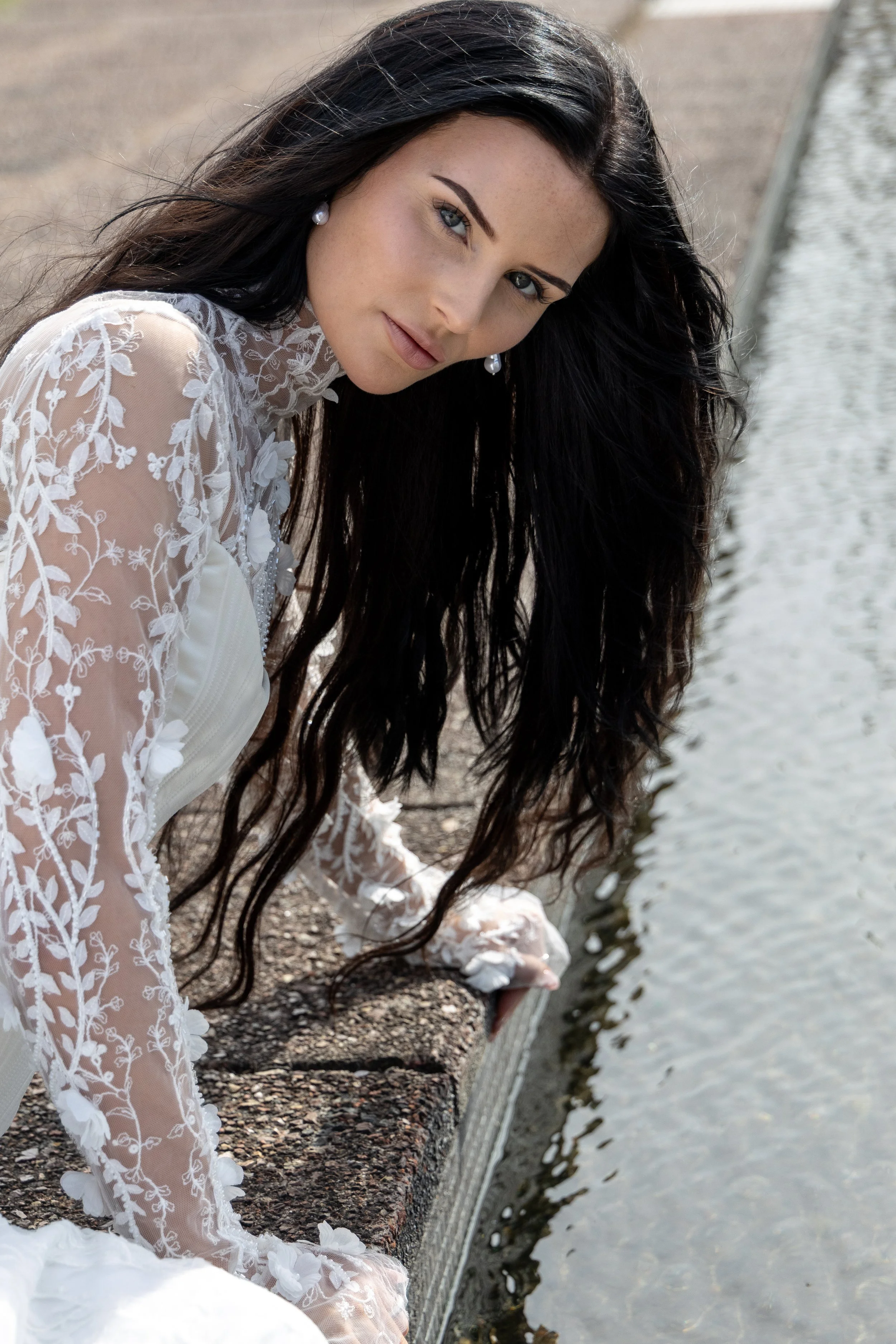 A woman with long black hair and pearl earrings, wearing a white lace dress, is sitting near the edge of a body of water with her hand dipping into the water.
