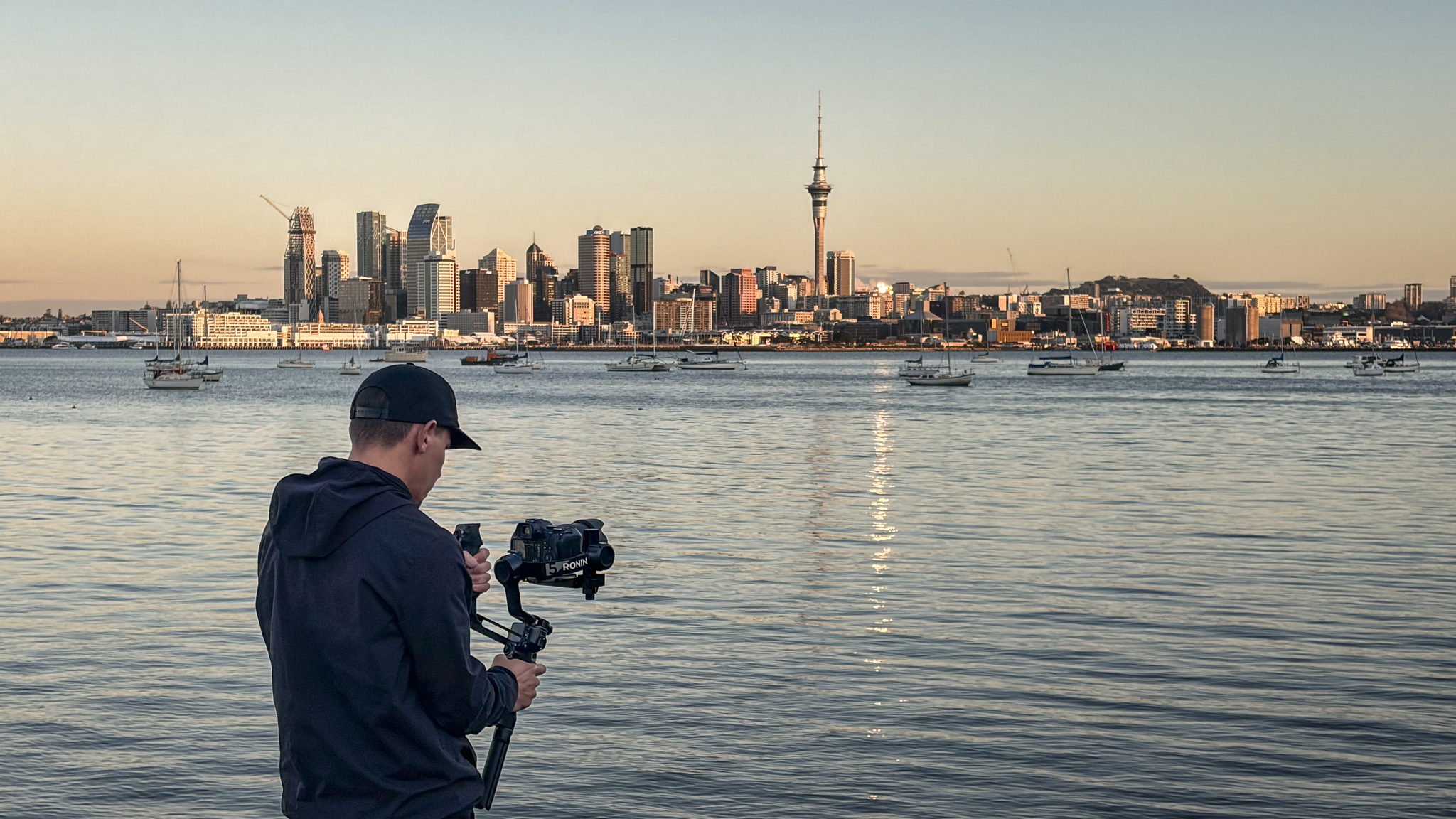 A person in a black jacket and cap operating a camera on a tripod by the water, with a city skyline and a tall tower in the background during sunset.