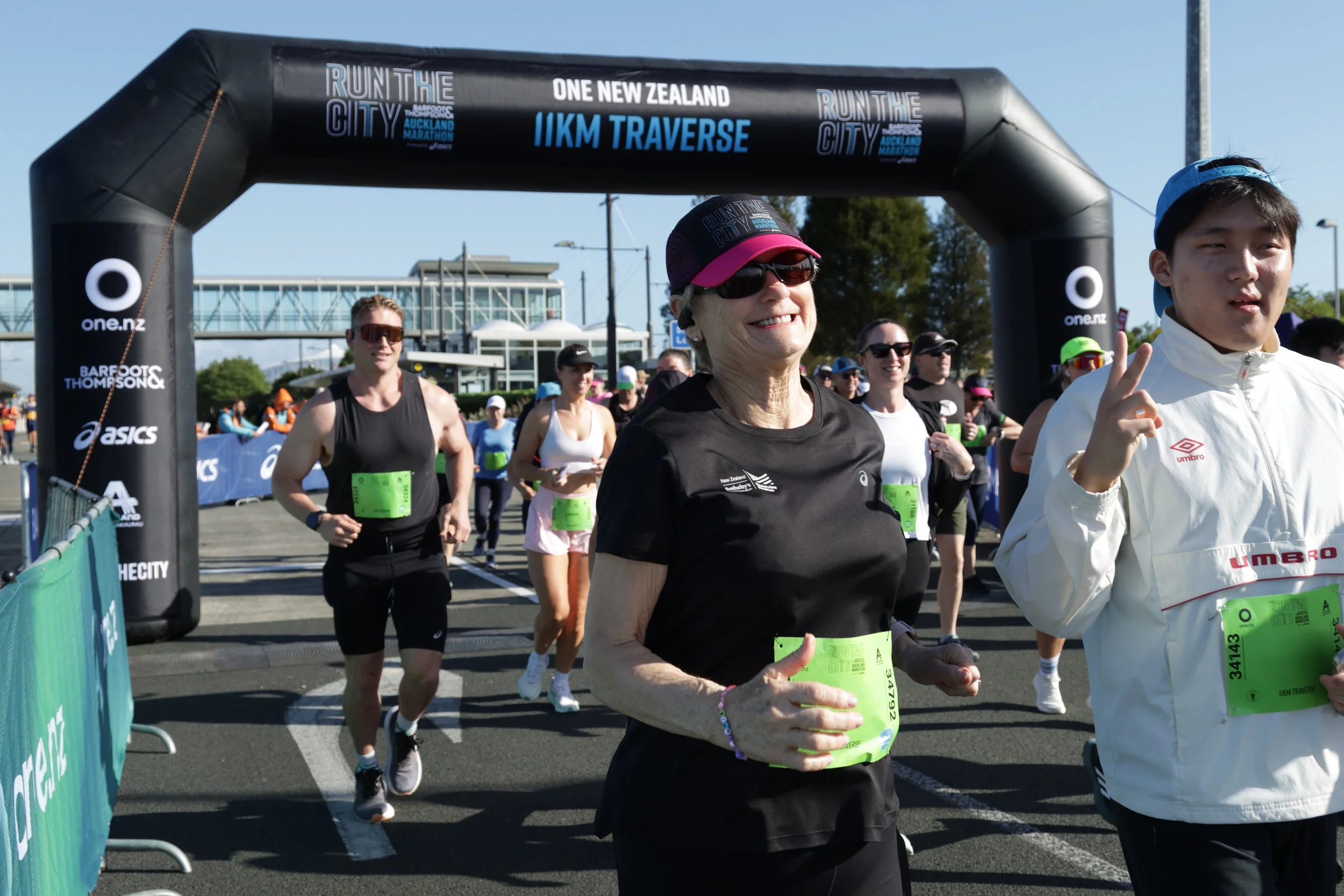 Multiple runners participating in a marathon, crossing the start or finish line under an inflatable arch that reads 'Run the City' and '1KM Traverse,' with onlookers and buildings in the background on a sunny day.