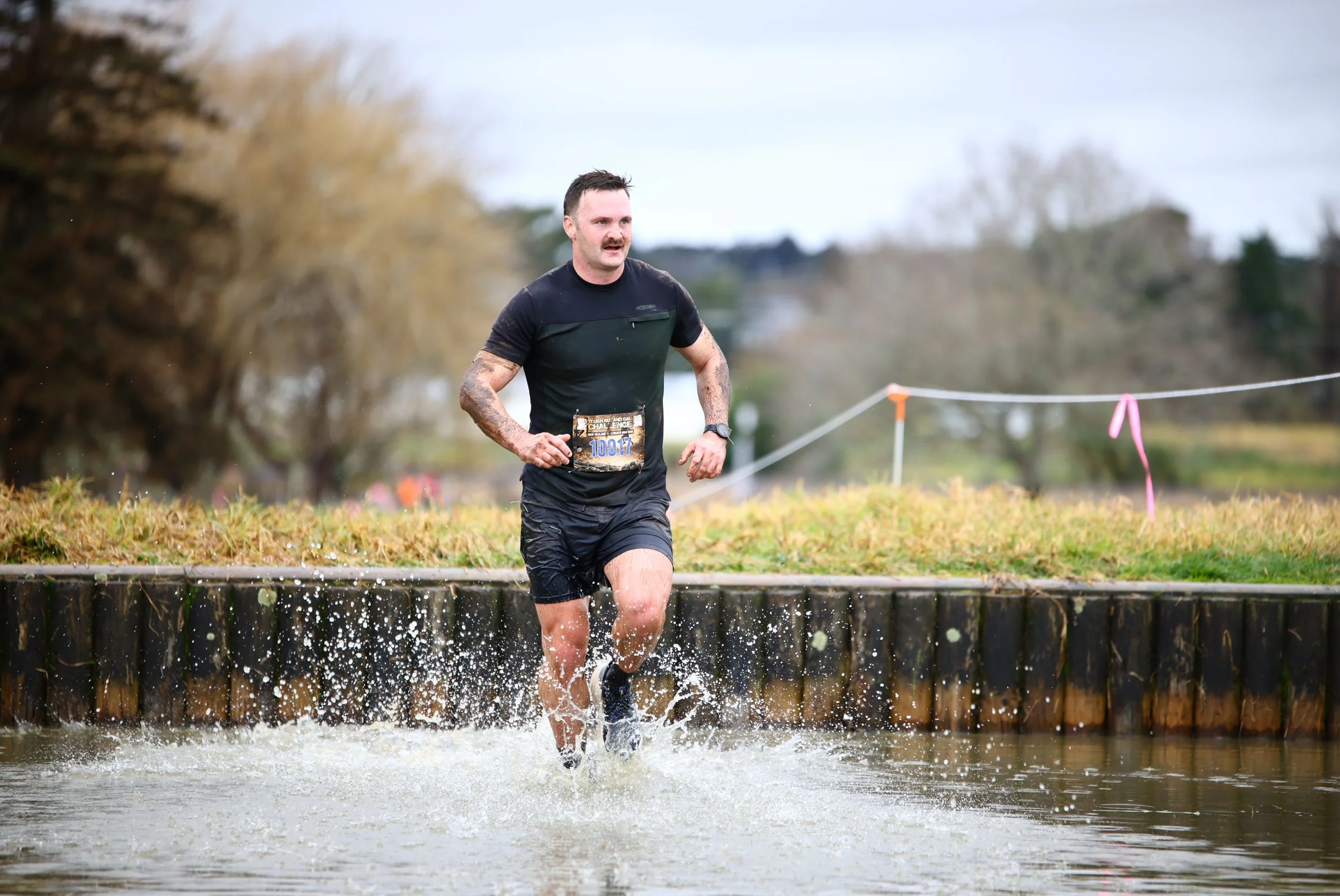 A man participating in a mud run or obstacle course race, running through water and mud, wearing athletic clothing and a race bib.