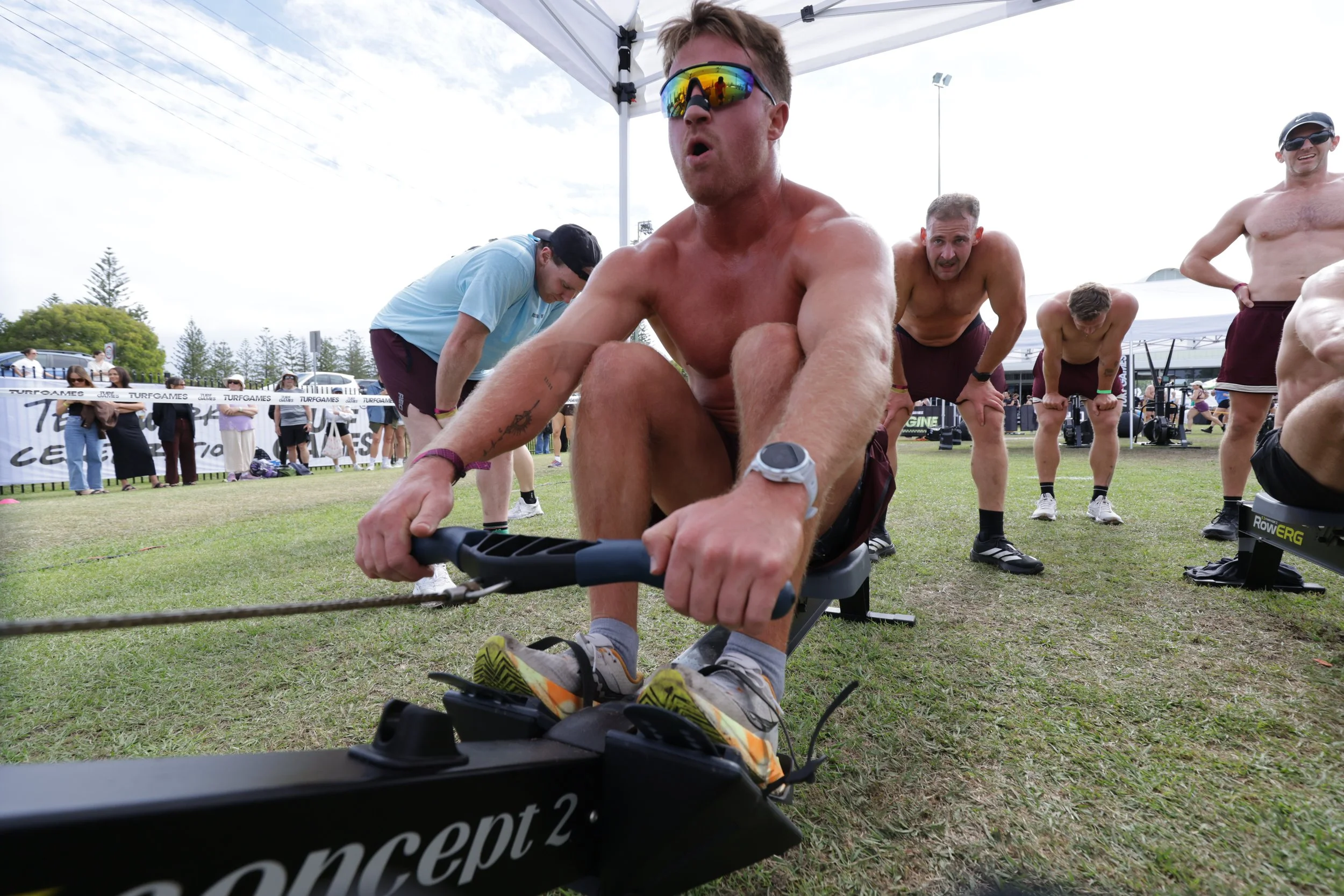 Group of shirtless men participating in a Strongman competition outdoors, with one man pulling a vehicle using a harness, while others watch and cheer.